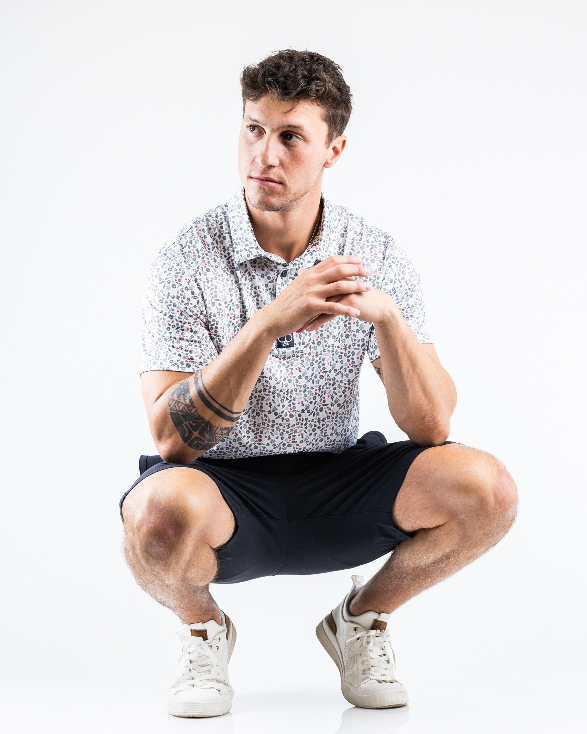 A man squatting in black performance shorts paired with a floral polo shirt, showcasing a stylish golf outfit.