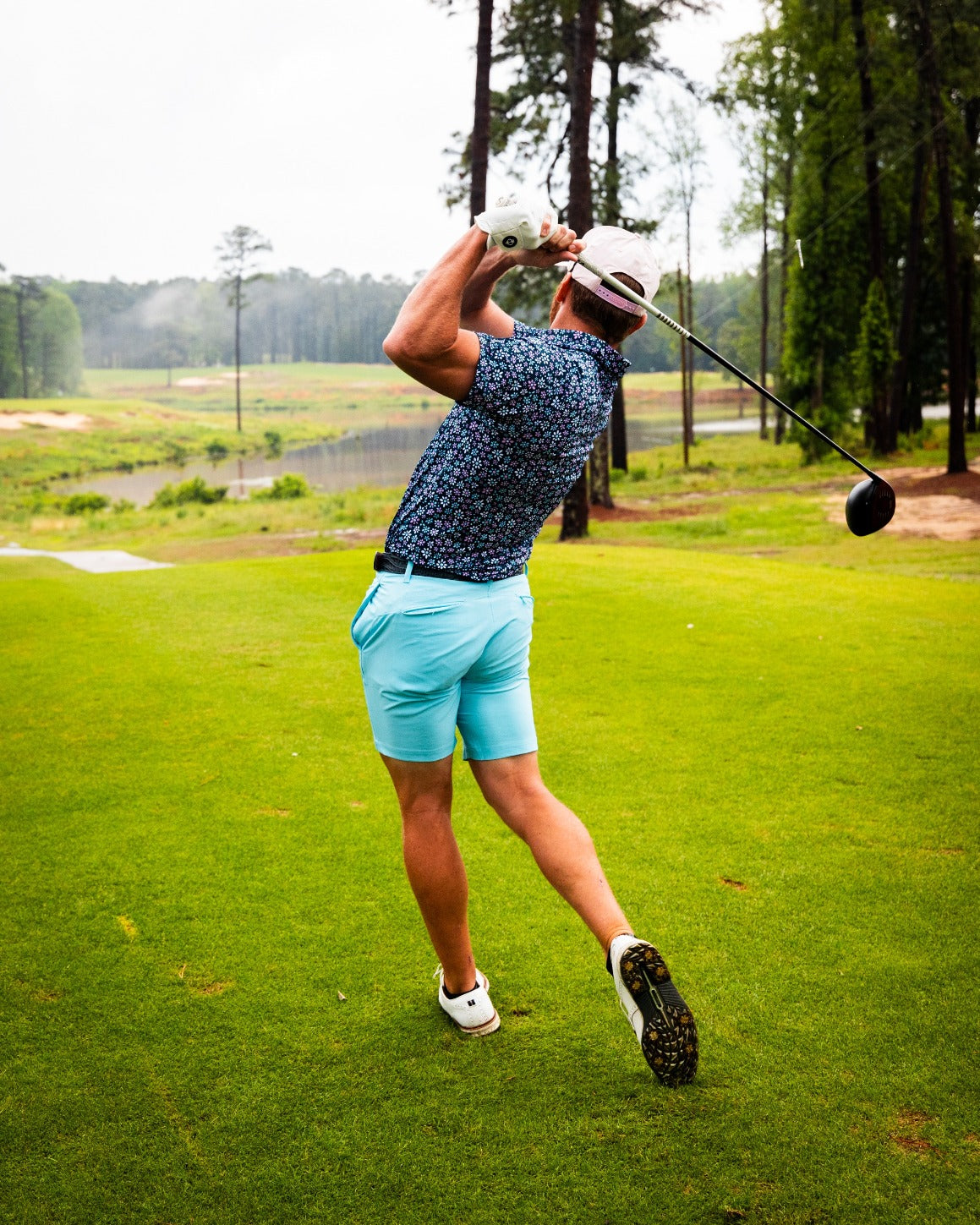 A golfer swings a driver on a lush course, wearing sky blue performance shorts and a floral shirt.