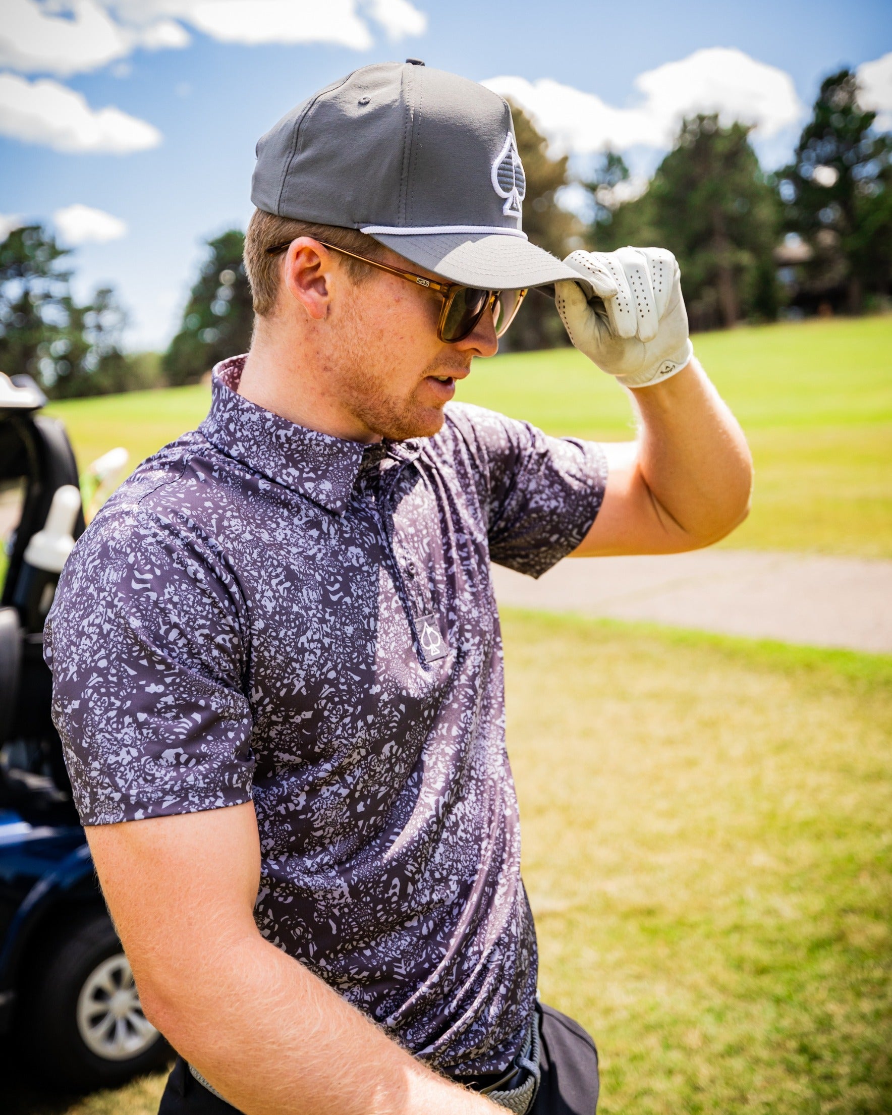 A golfer wearing a gray cap and sunglasses adjusts his hat while standing on the course, showcasing a stylish patterned polo shirt.