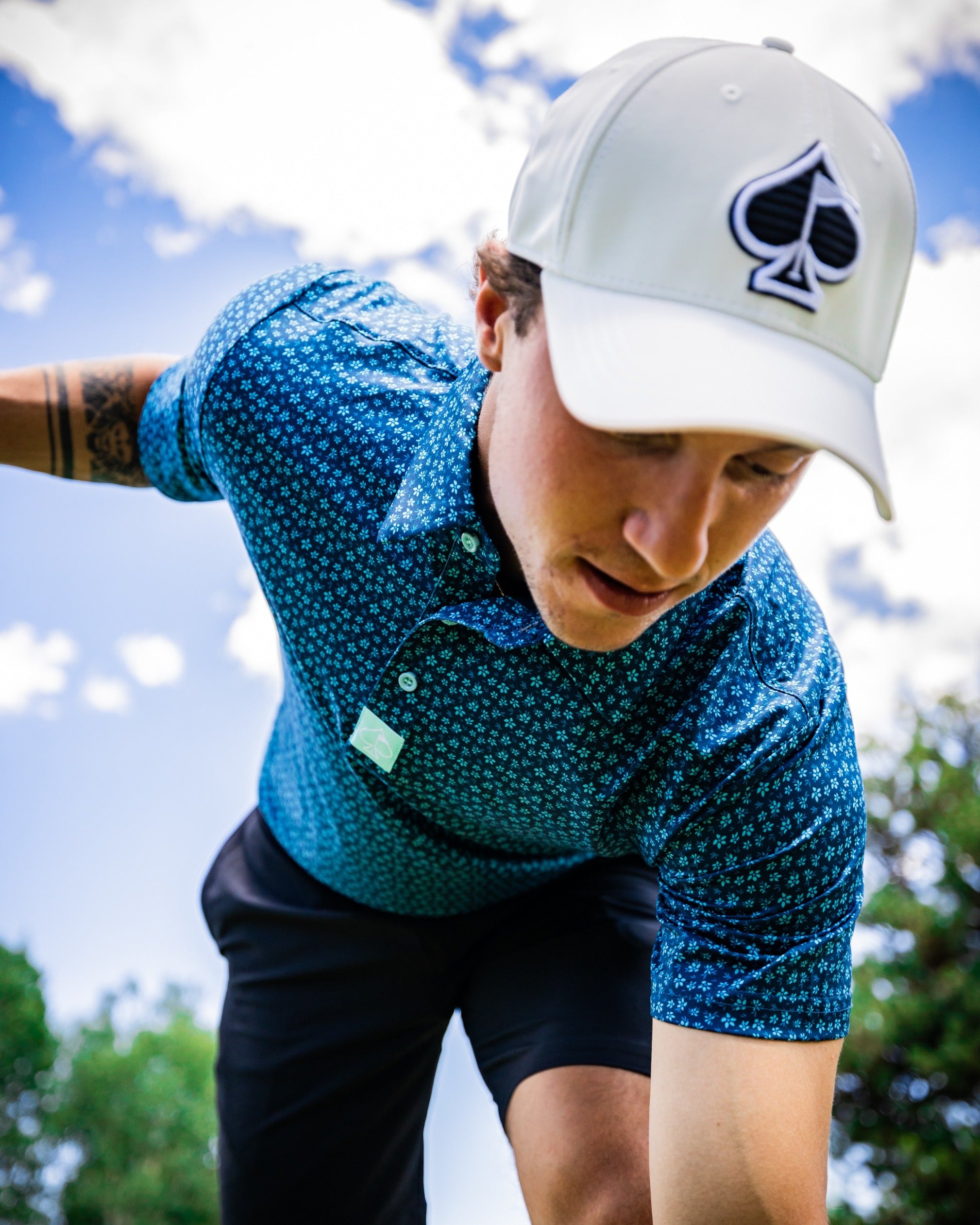 Man wearing a blue micro floral golf shirt and a light cap, focusing on his swing against a bright sky.