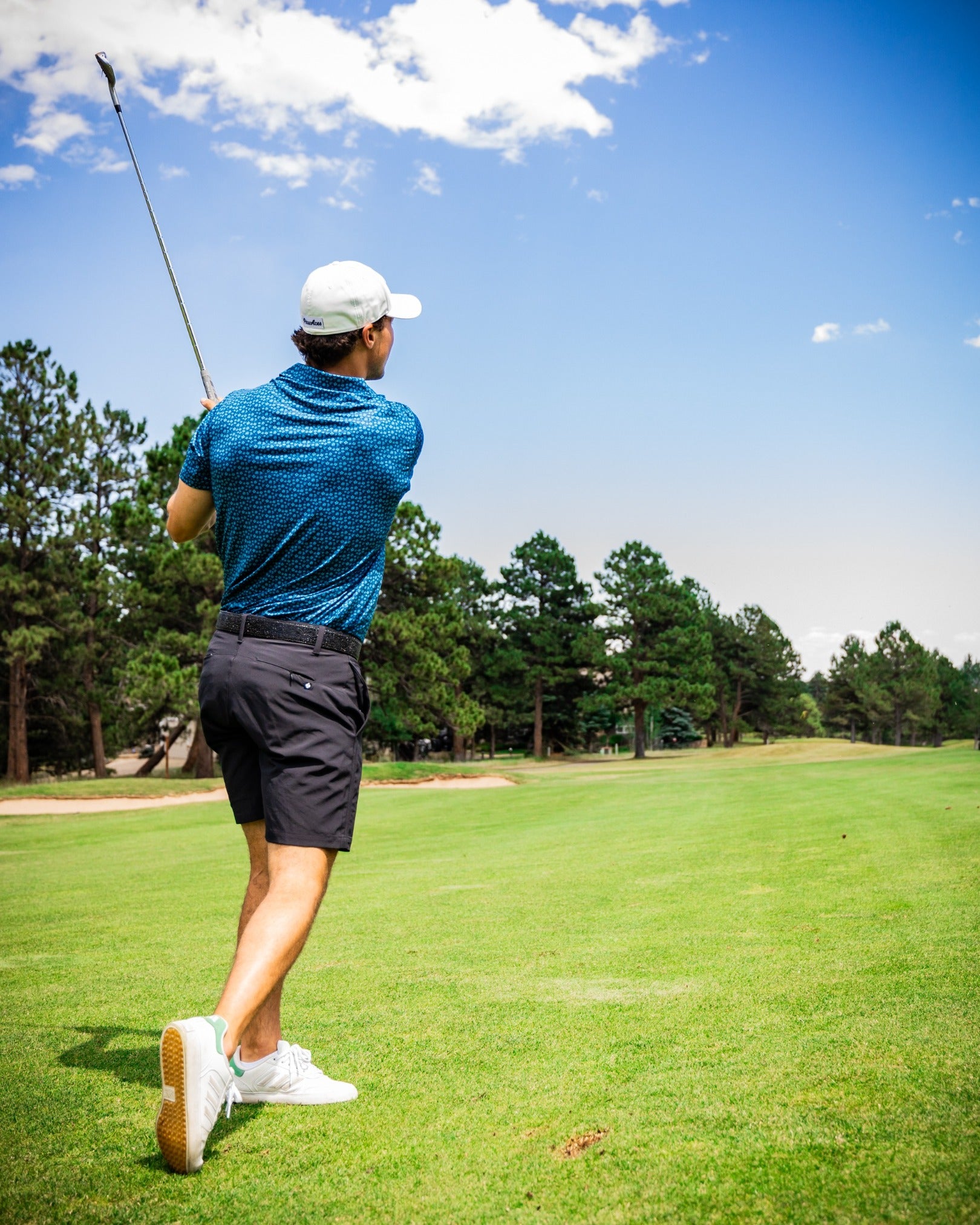 A golfer swings a club on a sunny course, wearing a blue micro floral polo and black shorts, showcasing bold golf style.