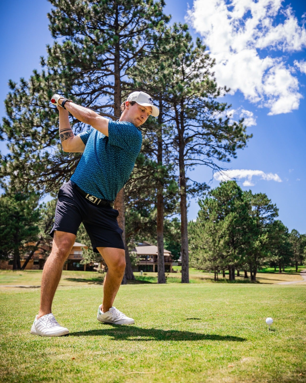 A golfer swings on the course, wearing a blue micro floral polo shirt and black shorts, surrounded by trees and clear skies.