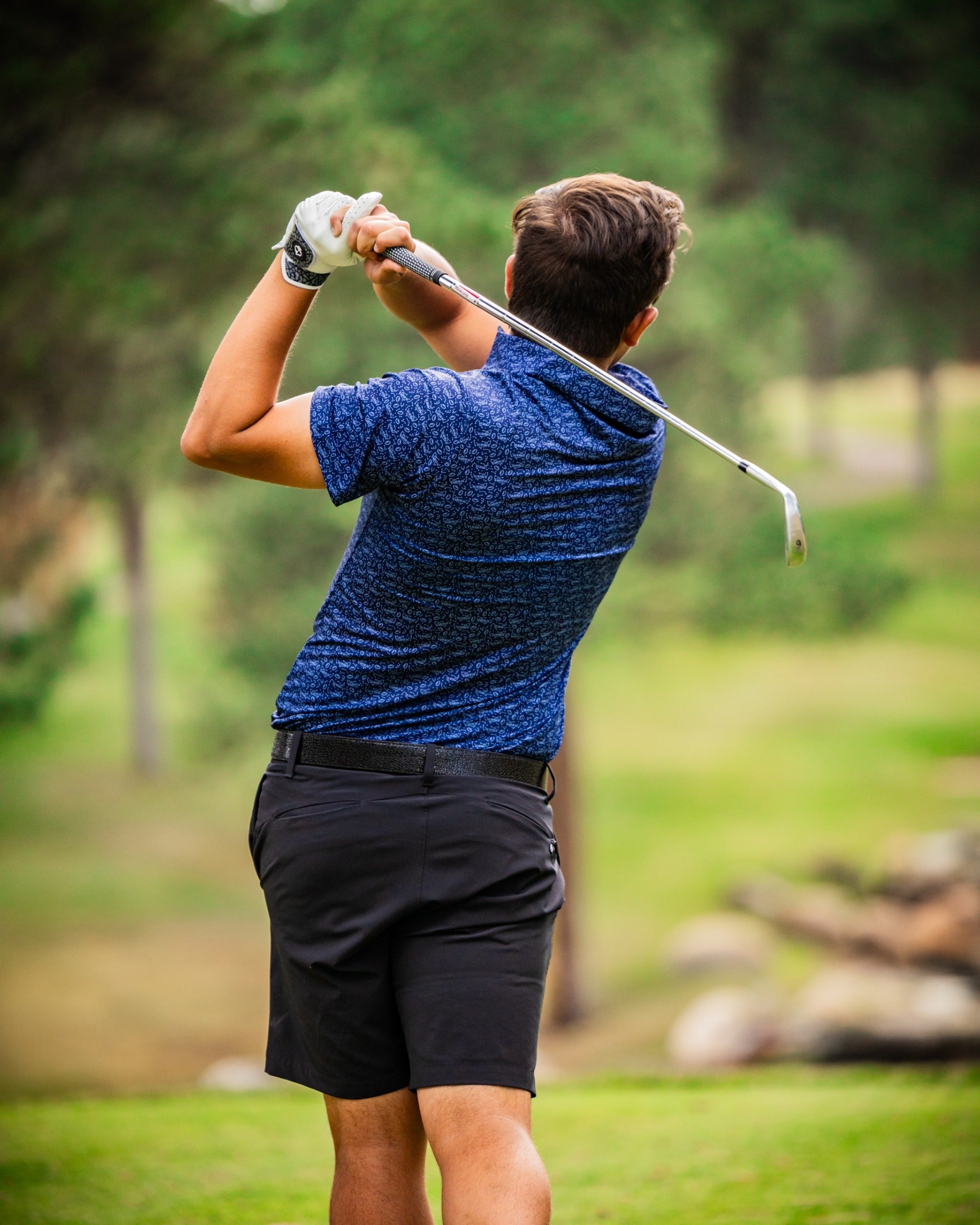 A golfer in a blue patterned polo and black shorts swings a club on a lush green course, showcasing athletic style.