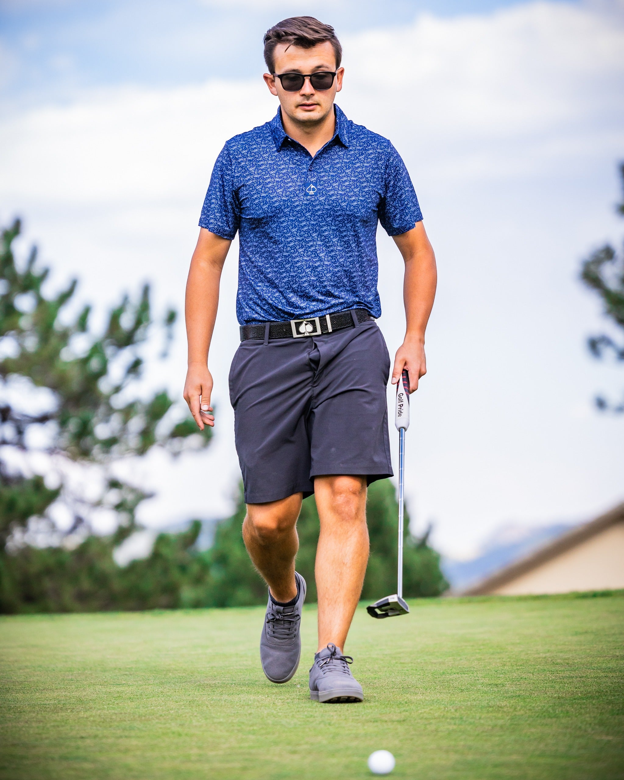 A golfer in a blue patterned polo and black shorts walks confidently on the green, holding a putter.