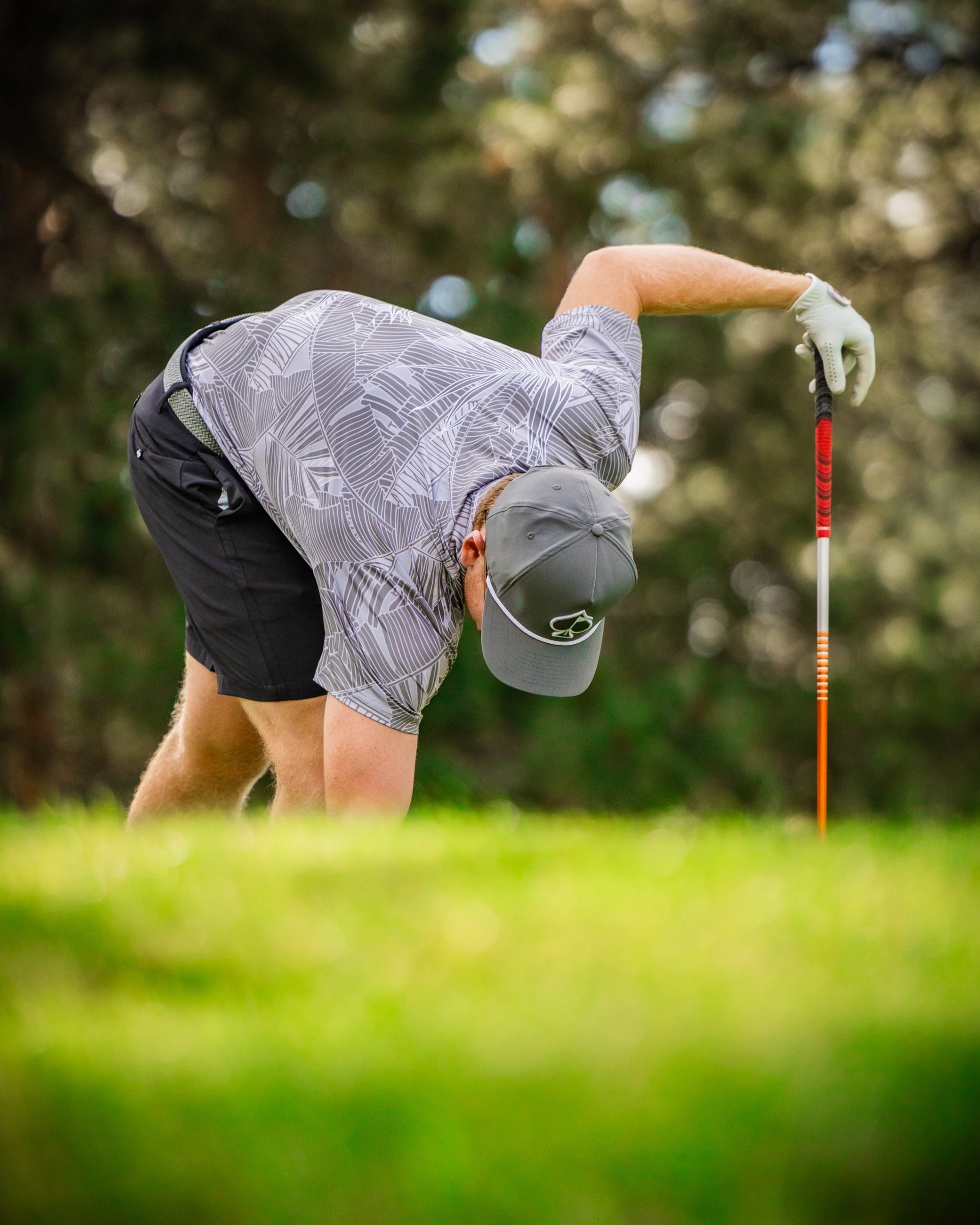 A golfer in a stylish palm-patterned shirt and shorts bends down to pick up a golf tee on a sunny course.