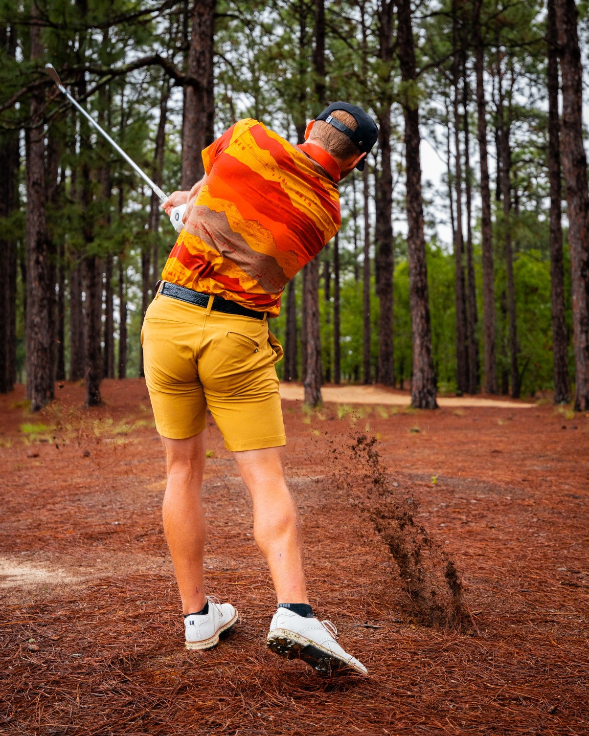A golfer swings a club in a vibrant Yellowstone Montana Sunset shirt, surrounded by tall trees and earthy tones.