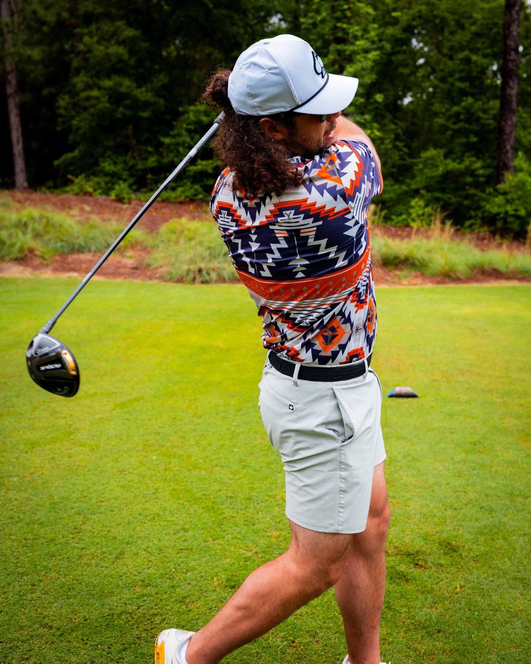A golfer swings a driver on the course, wearing a colorful Yellowstone Rustic Ranch shirt and light shorts.