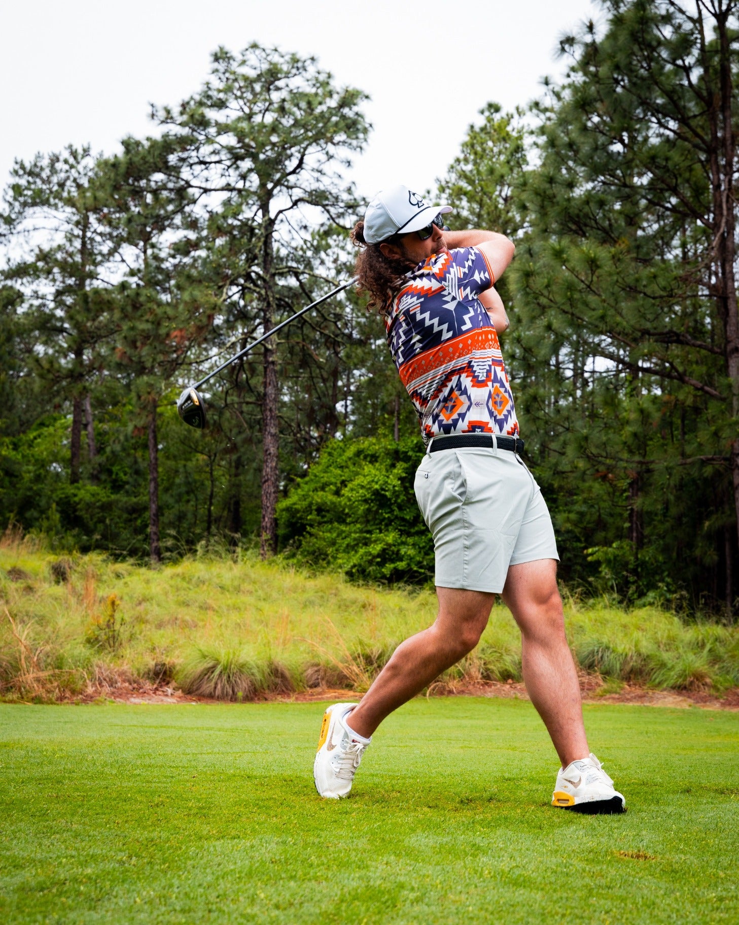 A golfer swings a driver on a lush course, wearing a colorful Yellowstone Rustic Ranch polo and light shorts.