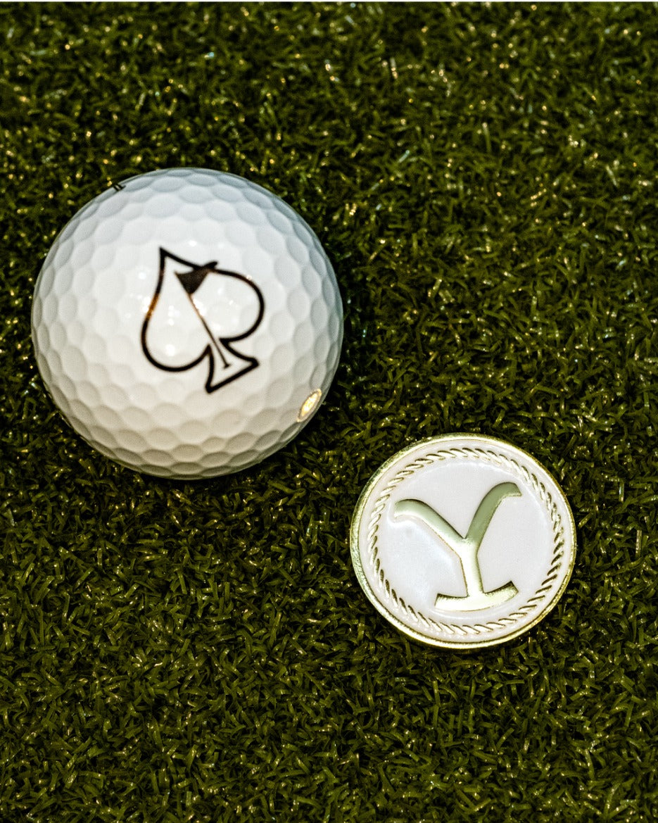 Yellowstone ball marker beside a golf ball featuring a spade logo, set against a green turf background.