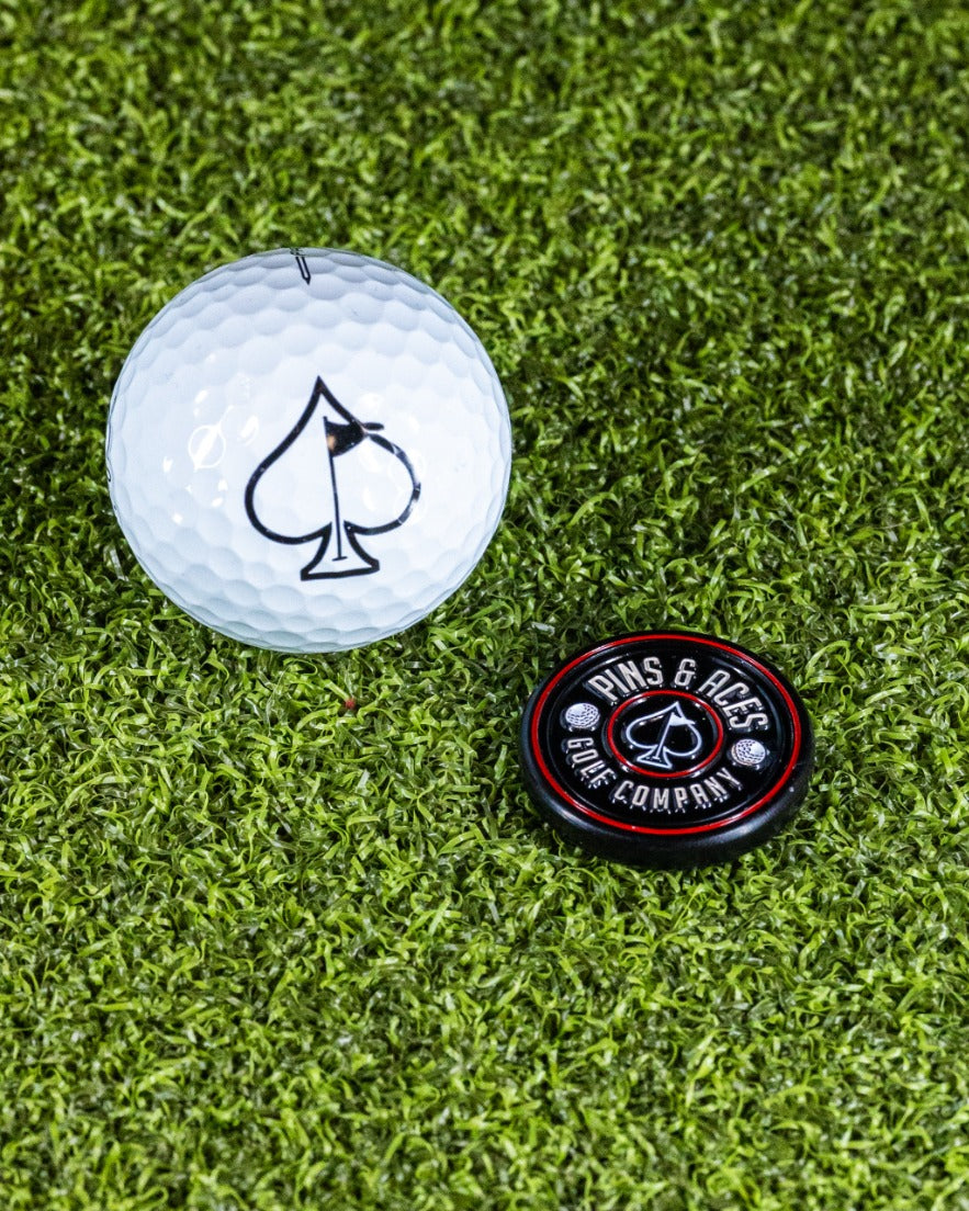Black Pop Out Medallion Ball Marker next to a white golf ball featuring a spade logo on a green turf background.