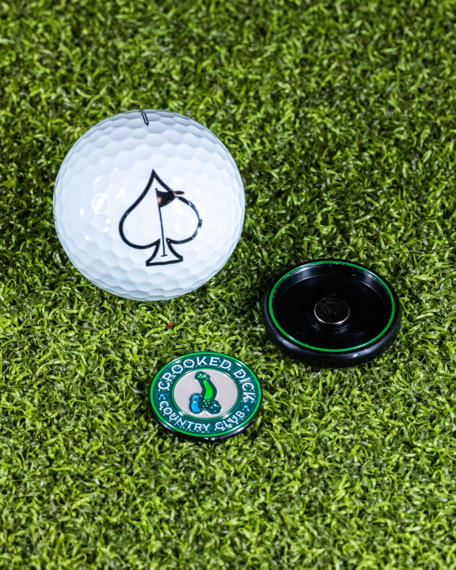 Pop Out Medallion Ball Marker featuring Crooked Dick design, alongside a golf ball with an ace of spades logo on grass.