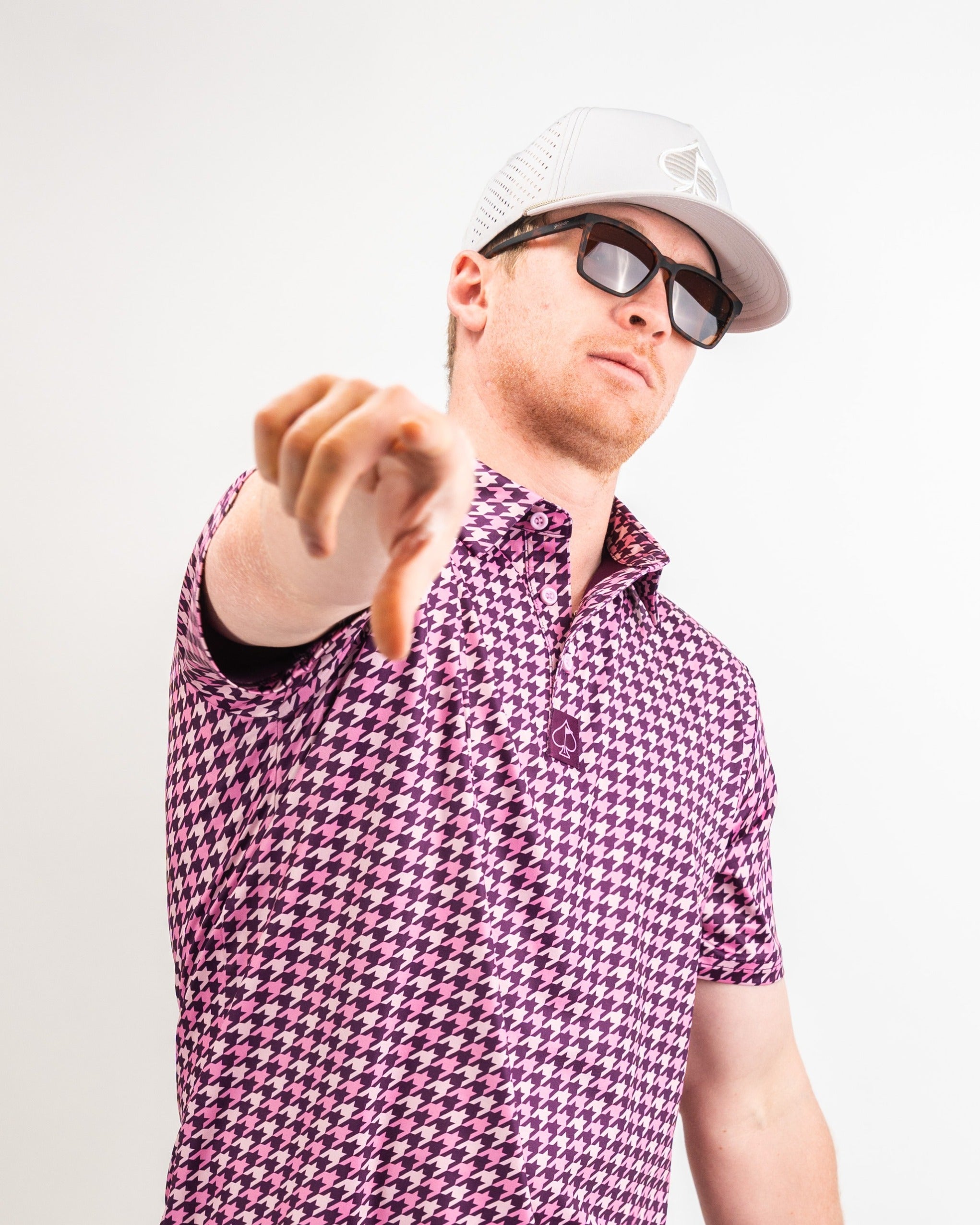 Bold houndstooth golf polo in raspberry pink, worn by a model in sunglasses and a cap, showcasing a stylish athletic look.