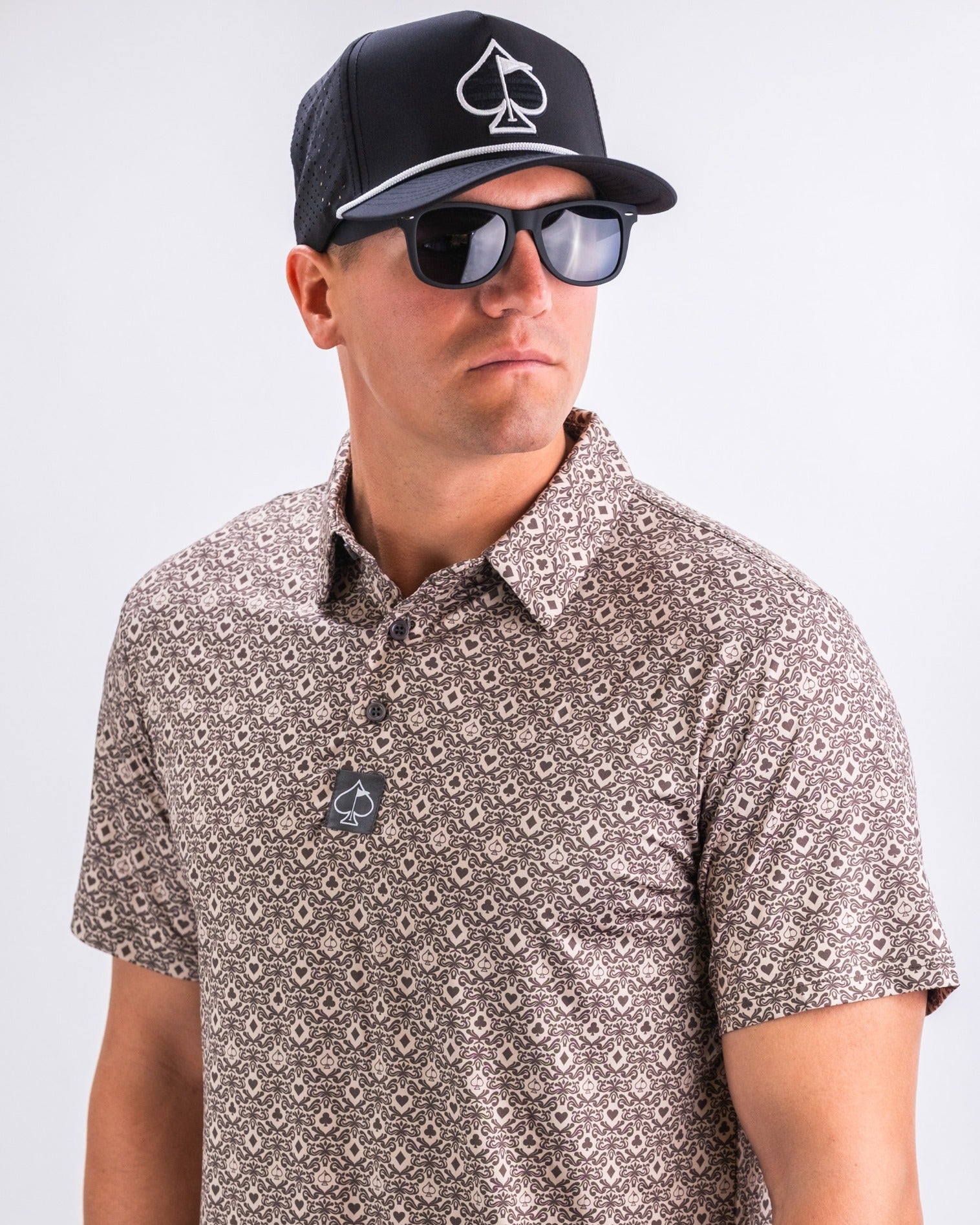 Man wearing a patterned golf polo shirt and a black cap with a spade logo, paired with sunglasses for a stylish look.