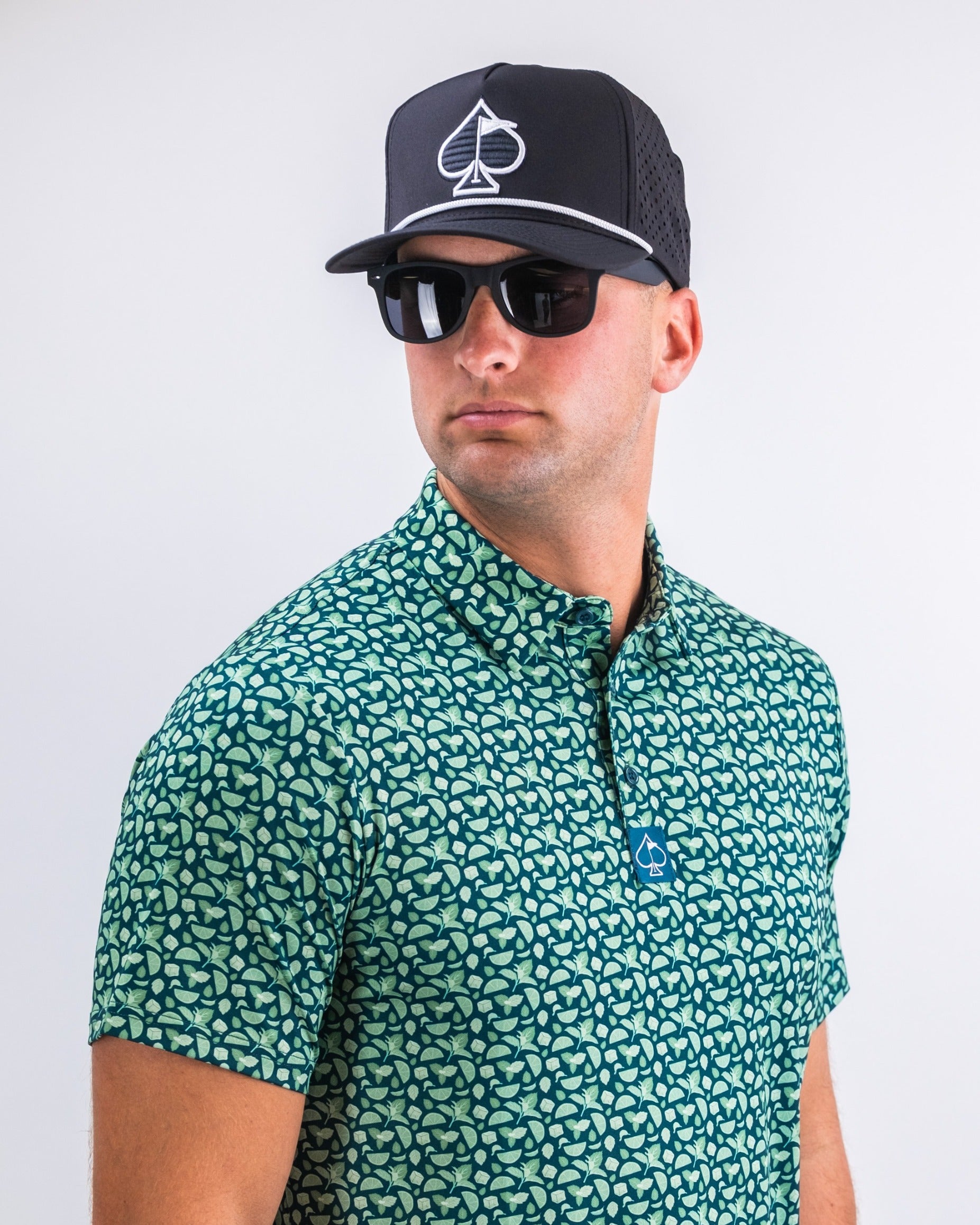 Man wearing a mint green patterned golf shirt and a black cap with a spade logo, accessorized with sunglasses.