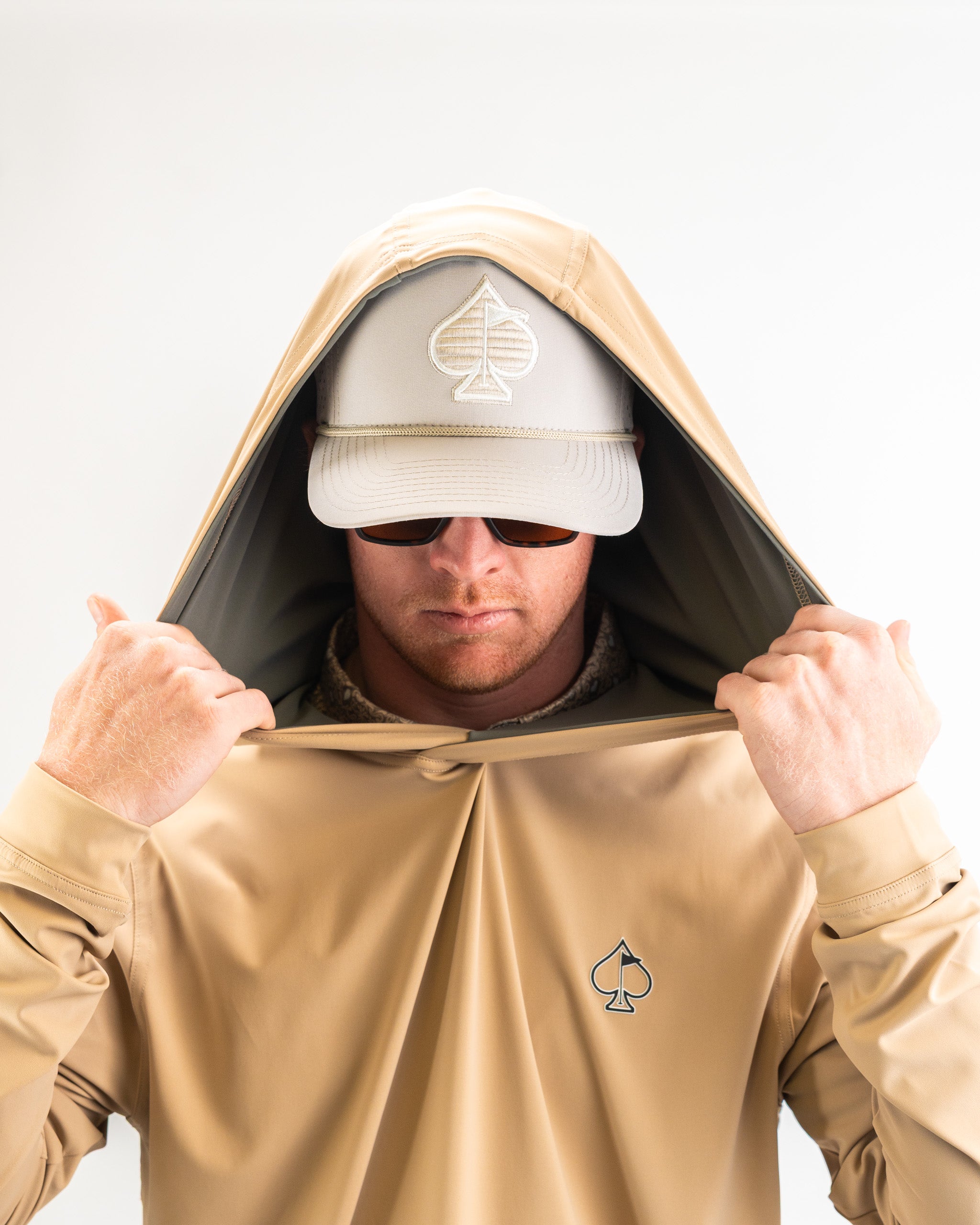 Man wearing a sand-colored performance golf hoodie with the hood up, showcasing a logo and a matching cap.