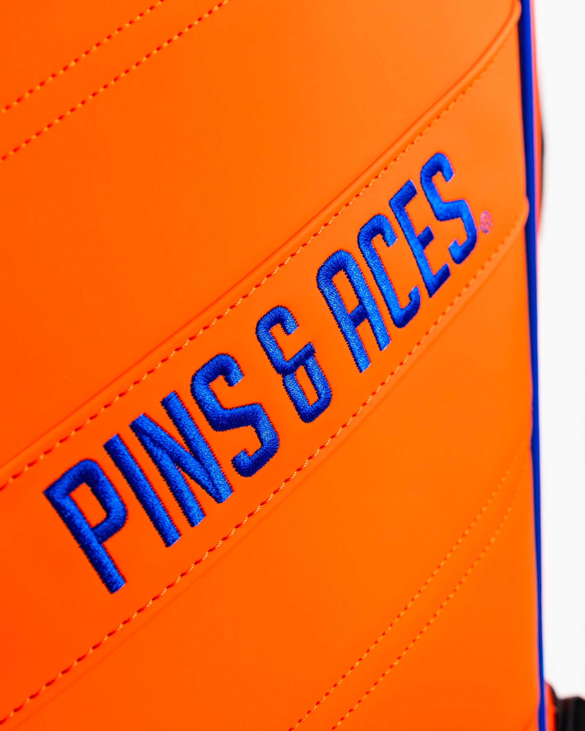 Vibrant orange golf bag featuring bold blue "Pins & Aces" branding, designed for stylish golfers.