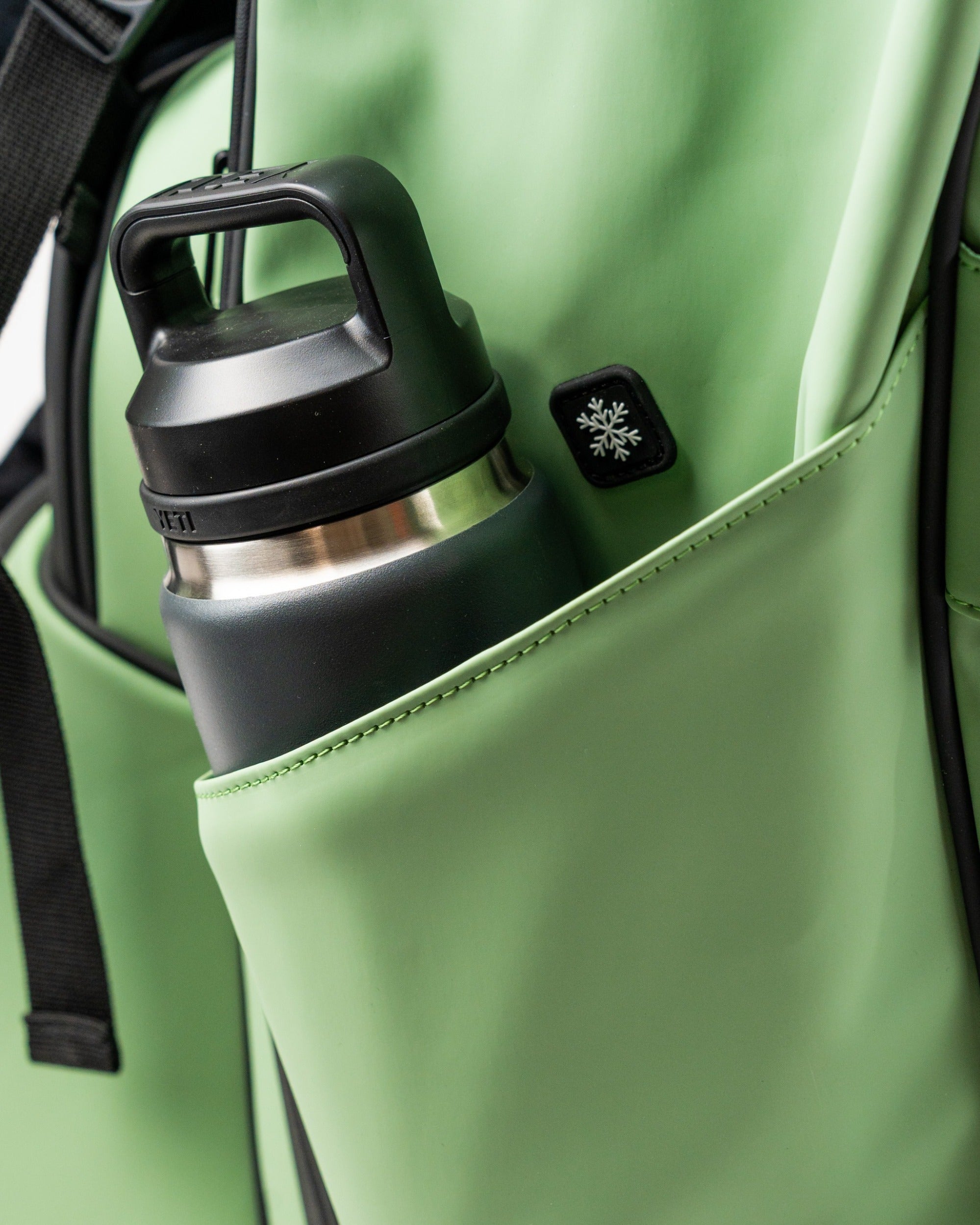 Close-up of a sage green golf bag featuring a side pocket with a black insulated water bottle and a small logo.