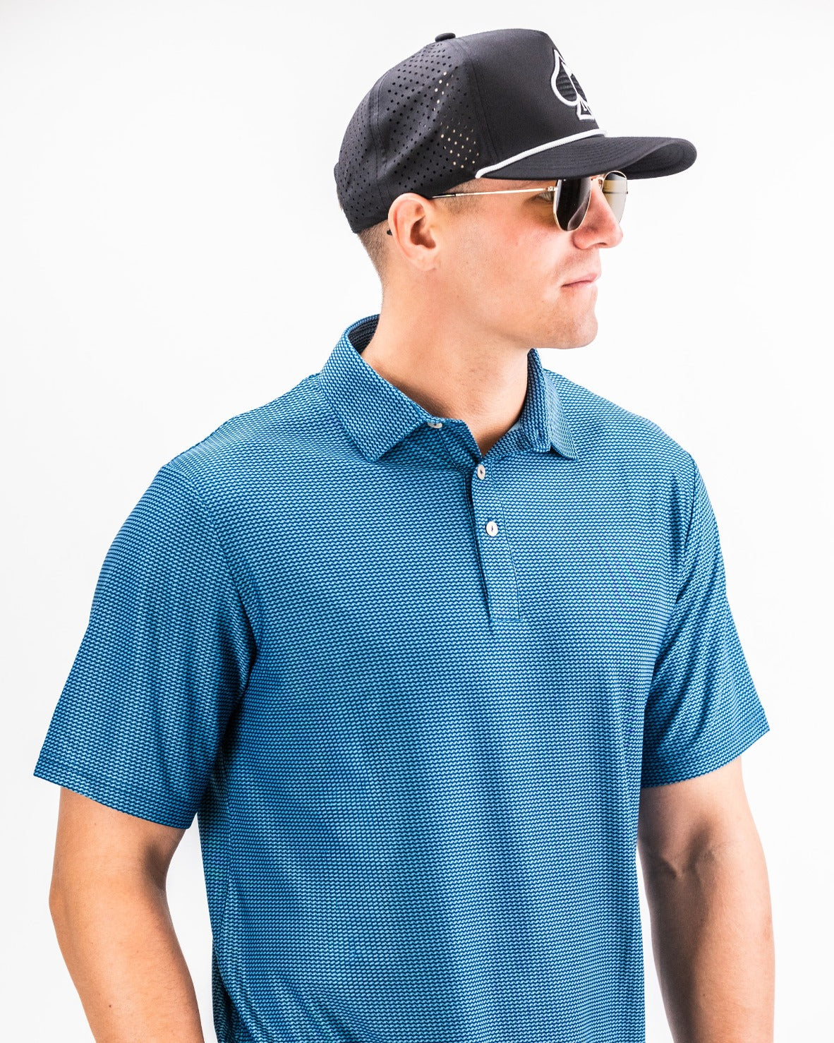 Model wearing a blue Player Preferred Polo with a subtle pattern, paired with a black cap and sunglasses, showcasing a stylish golf look.