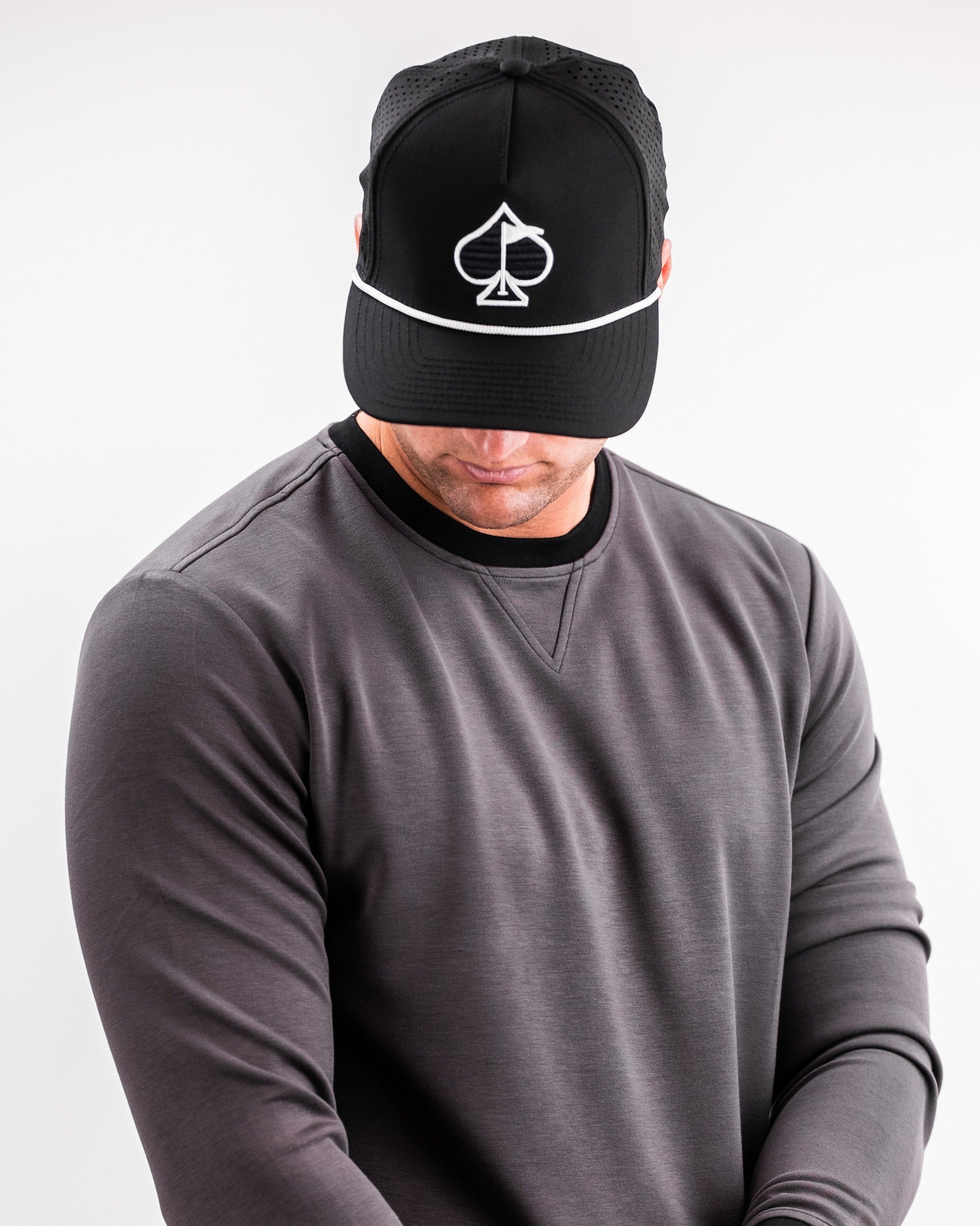 A man wearing a slate grey crewneck sweatshirt and a black cap with a spade logo, looking down thoughtfully.