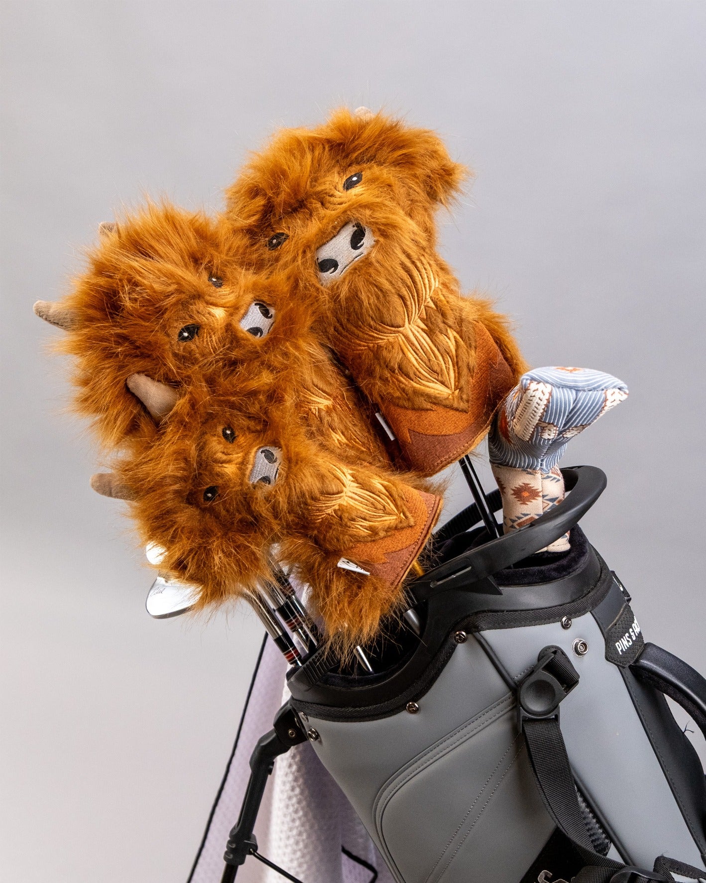 Highland cow hybrid golf headcovers featuring fluffy orange fur and playful designs, displayed on golf clubs in a bag.