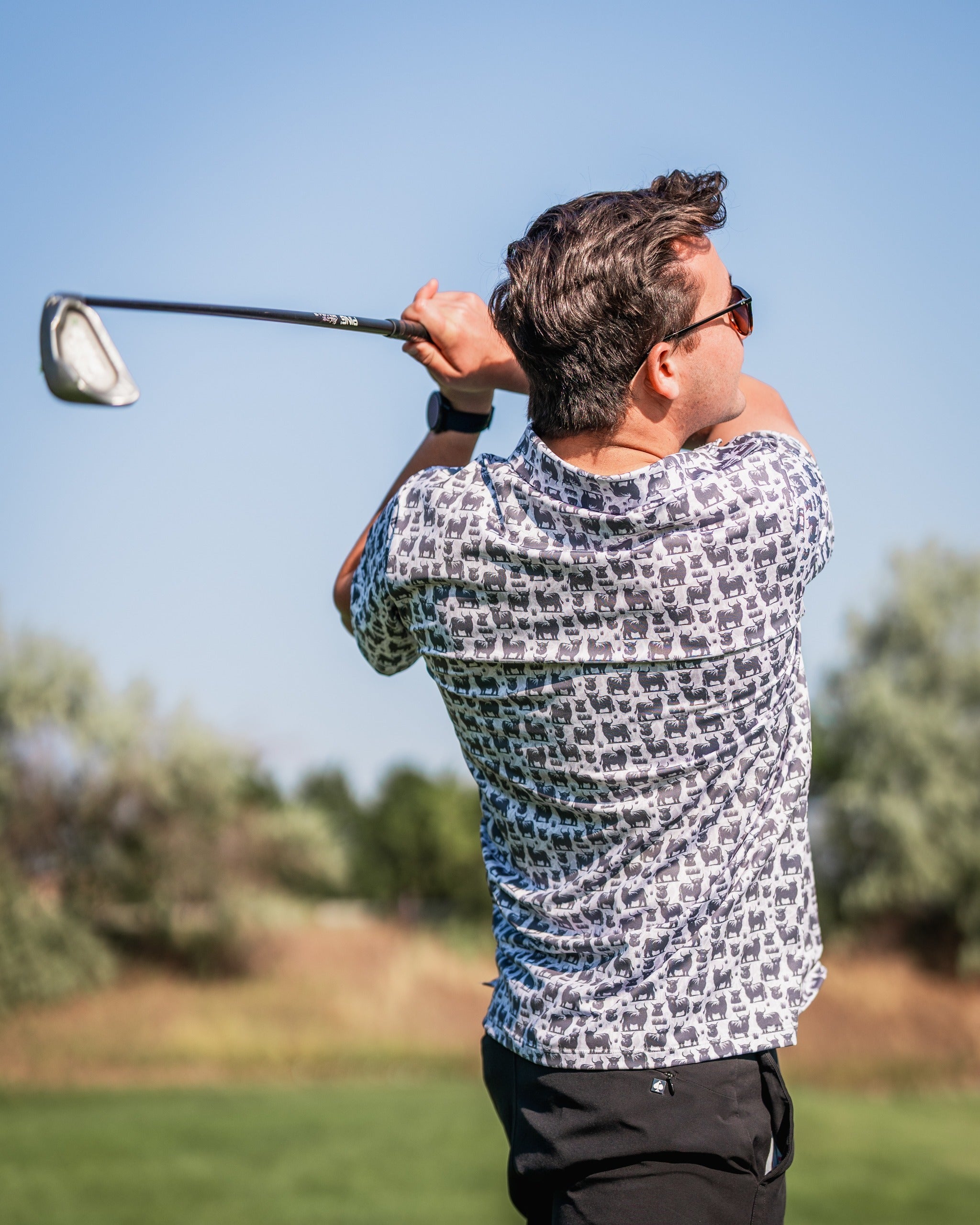 A golfer swings a club while wearing a Highland Cow patterned shirt on a sunny day at the golf course.