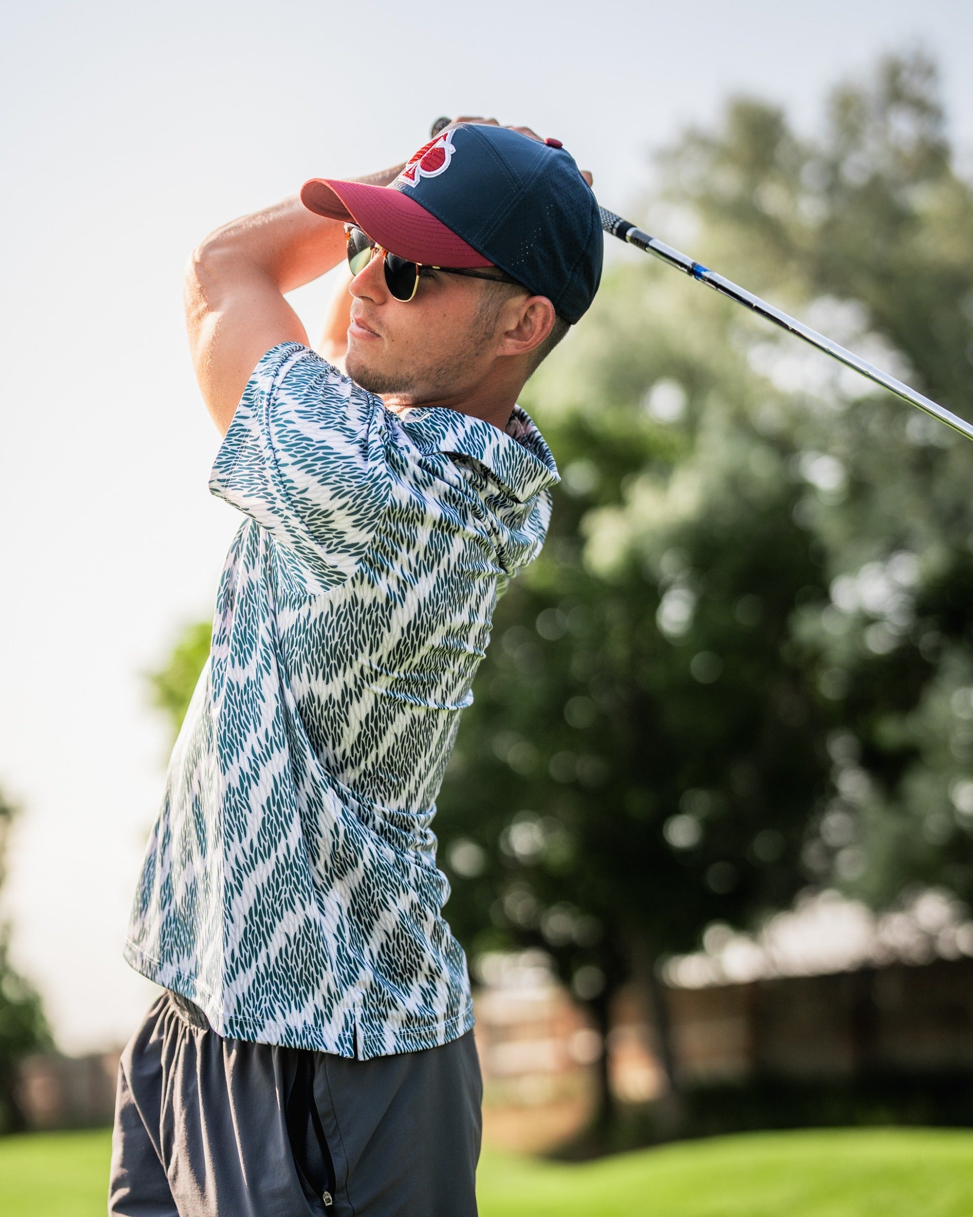 A golfer wearing a stylish patterned shirt and sunglasses swings a club on a sunny course, showcasing bold golf fashion.
