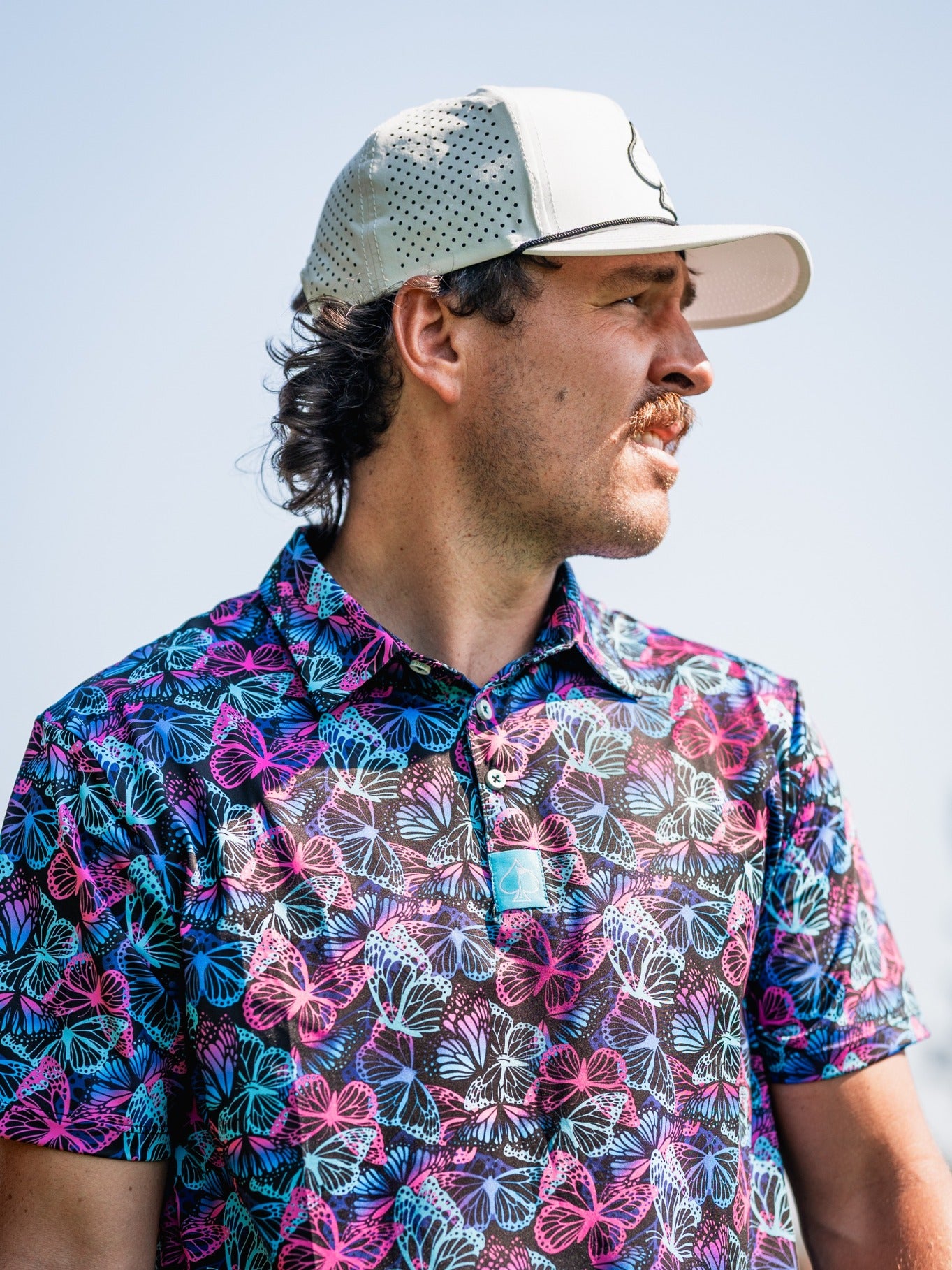 Man wearing a vibrant Electric Butterfly golf shirt featuring a colorful butterfly pattern, set against a clear sky.