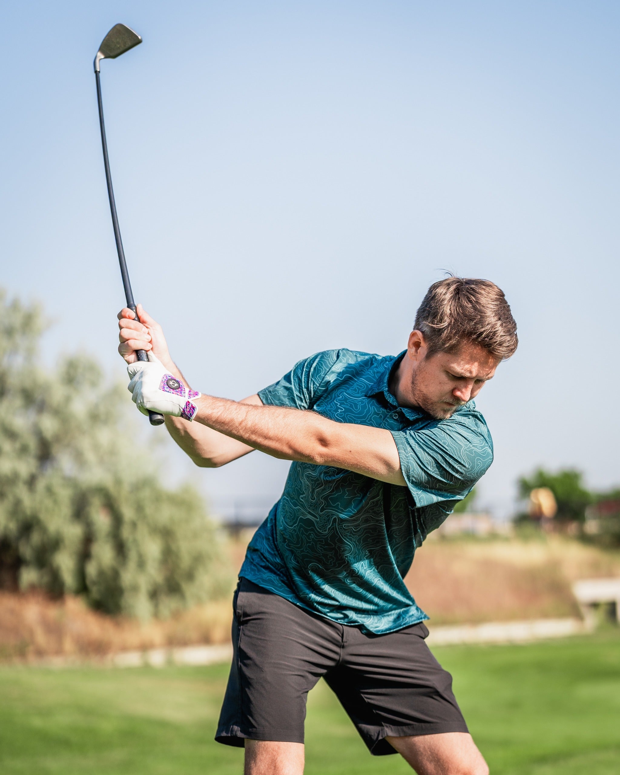 A golfer swings a club while wearing a teal Terrain polo shirt and black shorts on a sunny course.