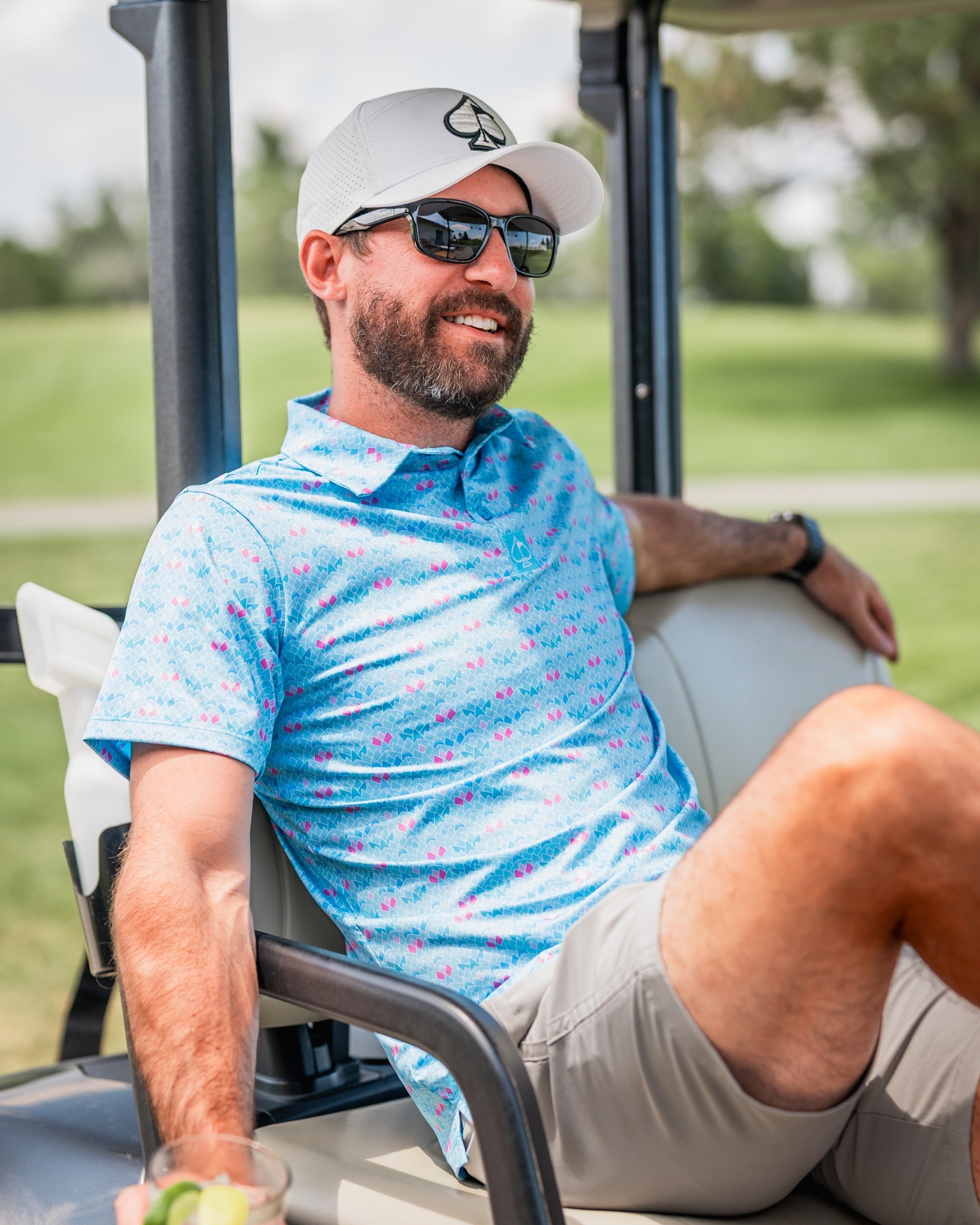 Man relaxing on a golf cart, wearing a blue floral polo shirt and sunglasses, enjoying a drink on the course.