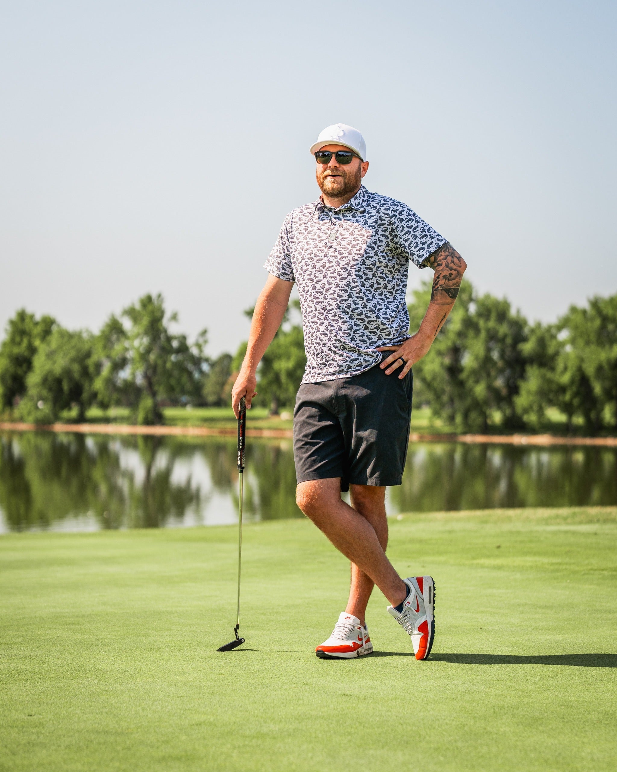 Man in a patterned golf shirt and shorts stands confidently on a golf course, holding a putter with a scenic backdrop.
