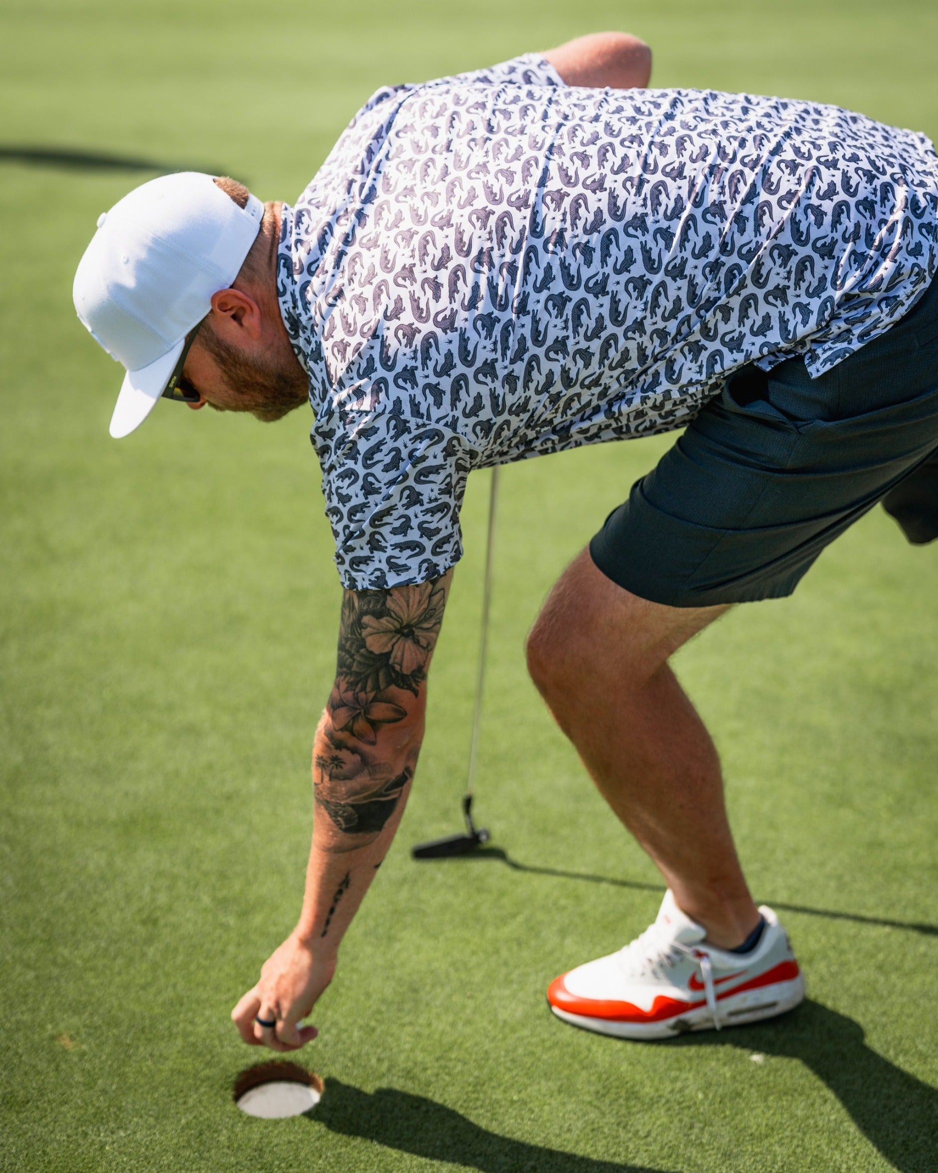 A golfer in a patterned shirt and shorts retrieves a golf ball from the hole on a green.