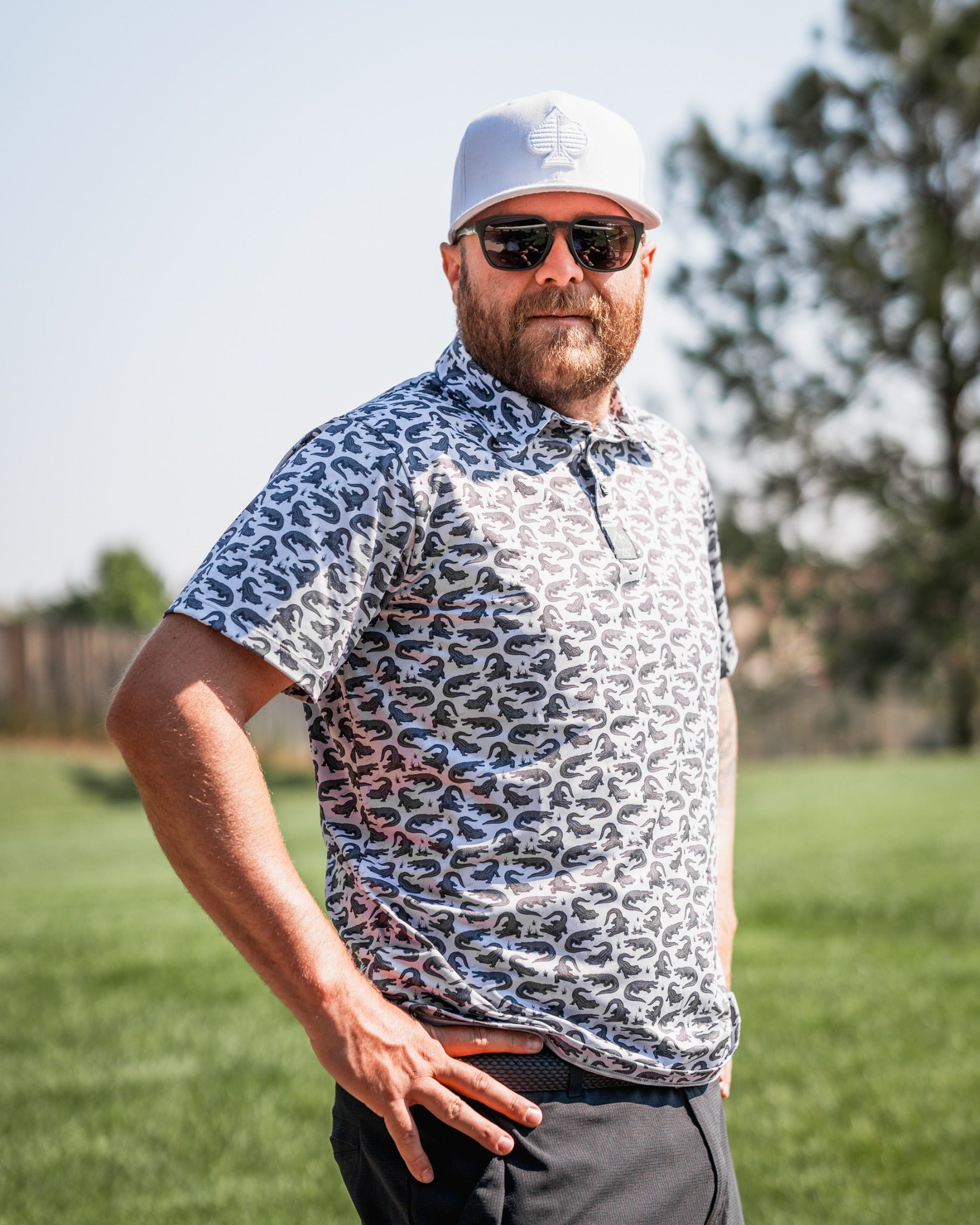 Stylish golfer wearing a Gator-patterned polo shirt, sunglasses, and a cap, posing on a sunny golf course.