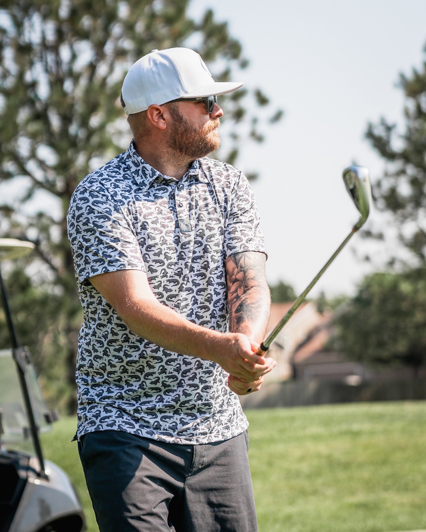 Man swinging a golf club wearing a Gator-patterned polo shirt on a sunny golf course.