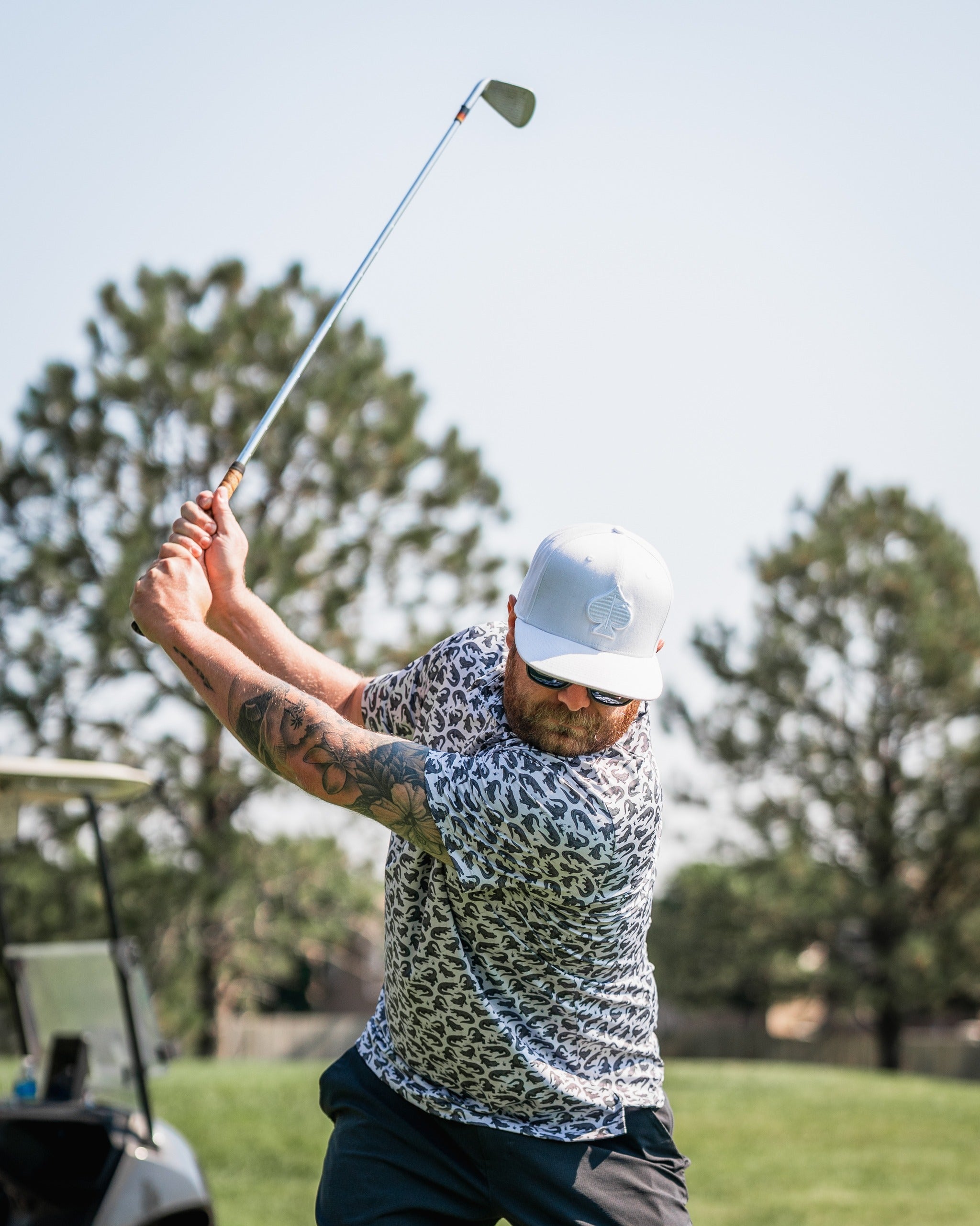 A golfer swings a club while wearing a bold, patterned shirt and a white cap, showcasing a stylish golf look.