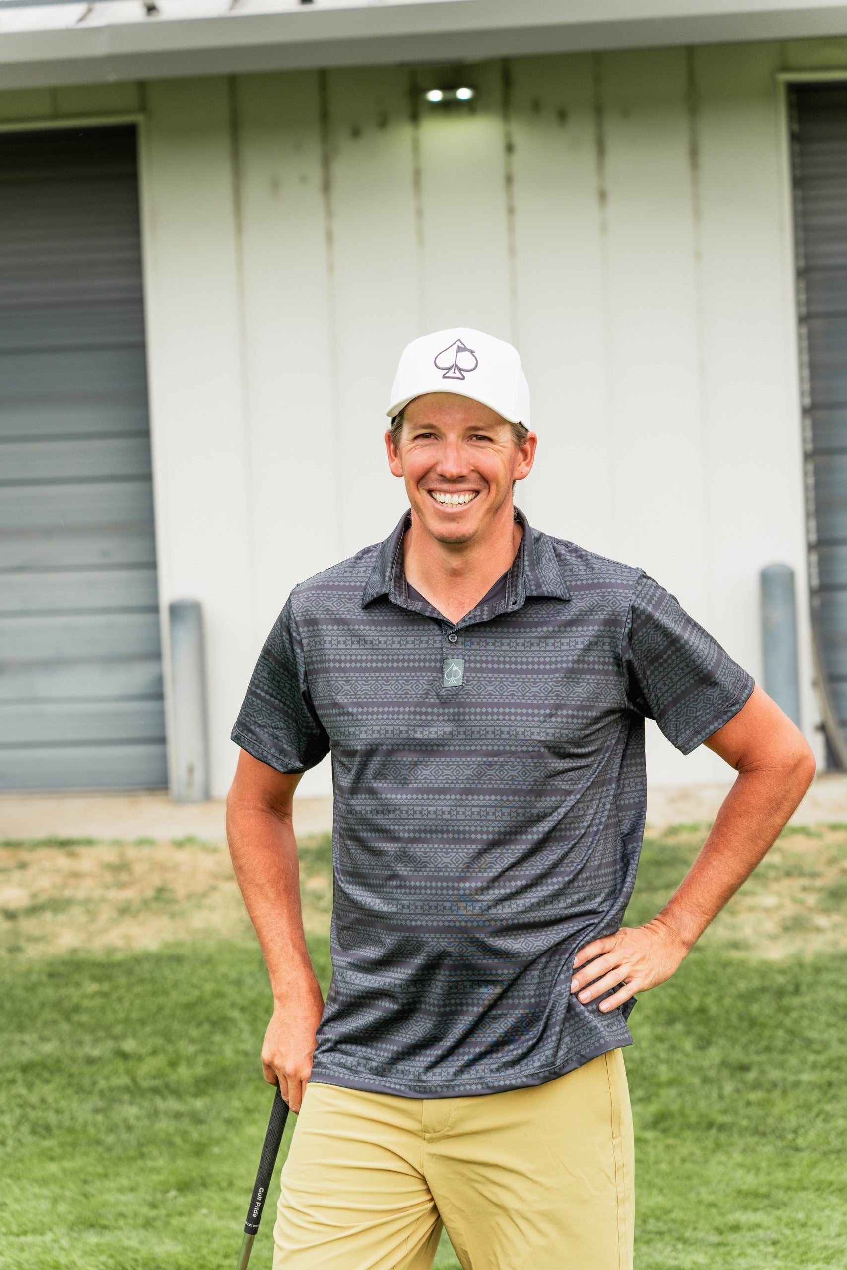 Man smiling on a golf course, wearing a patterned polo shirt and a white cap, holding a golf club.