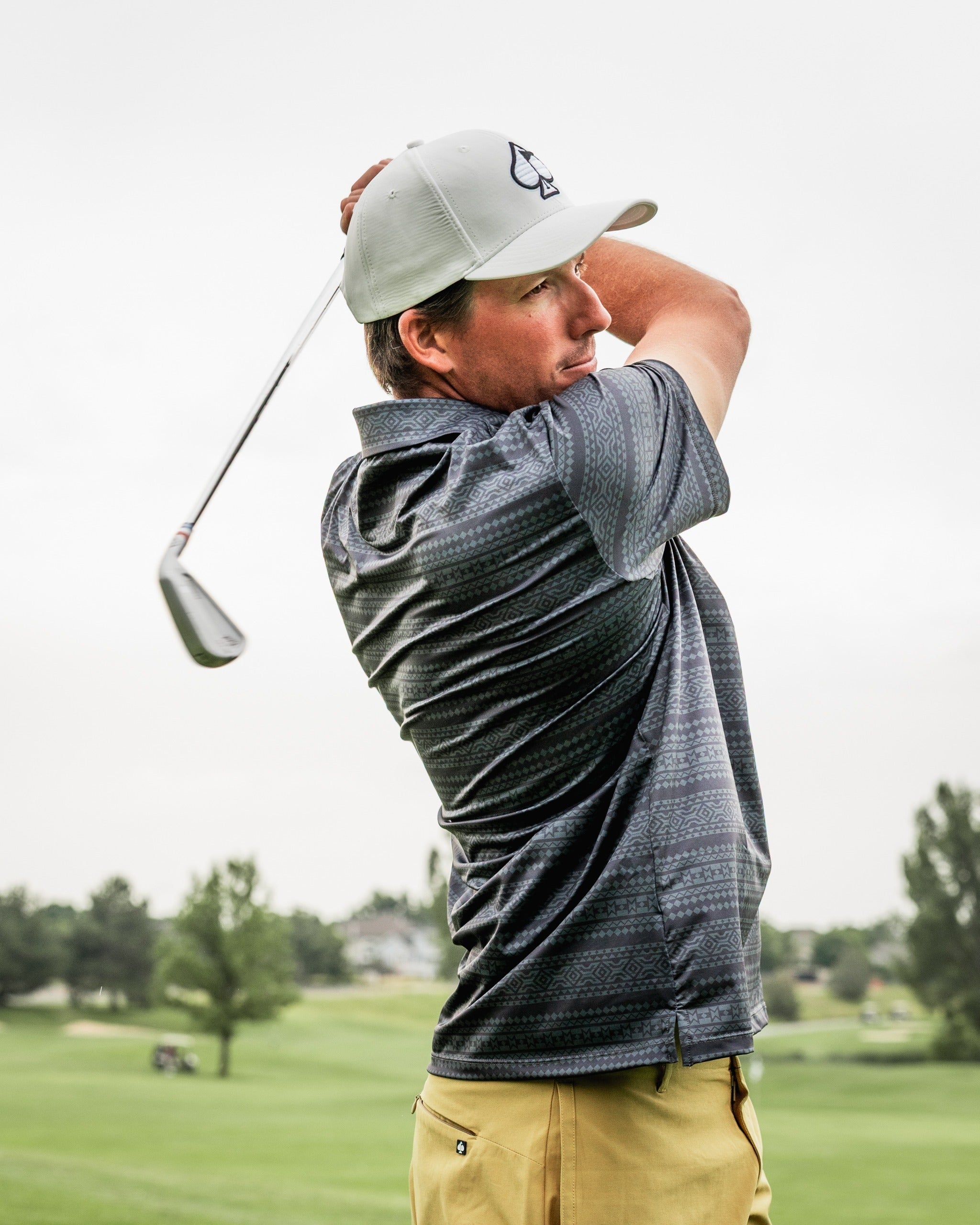 A golfer swings a club on the course, wearing a patterned gray shirt and a light cap, showcasing the Desert Geo apparel.