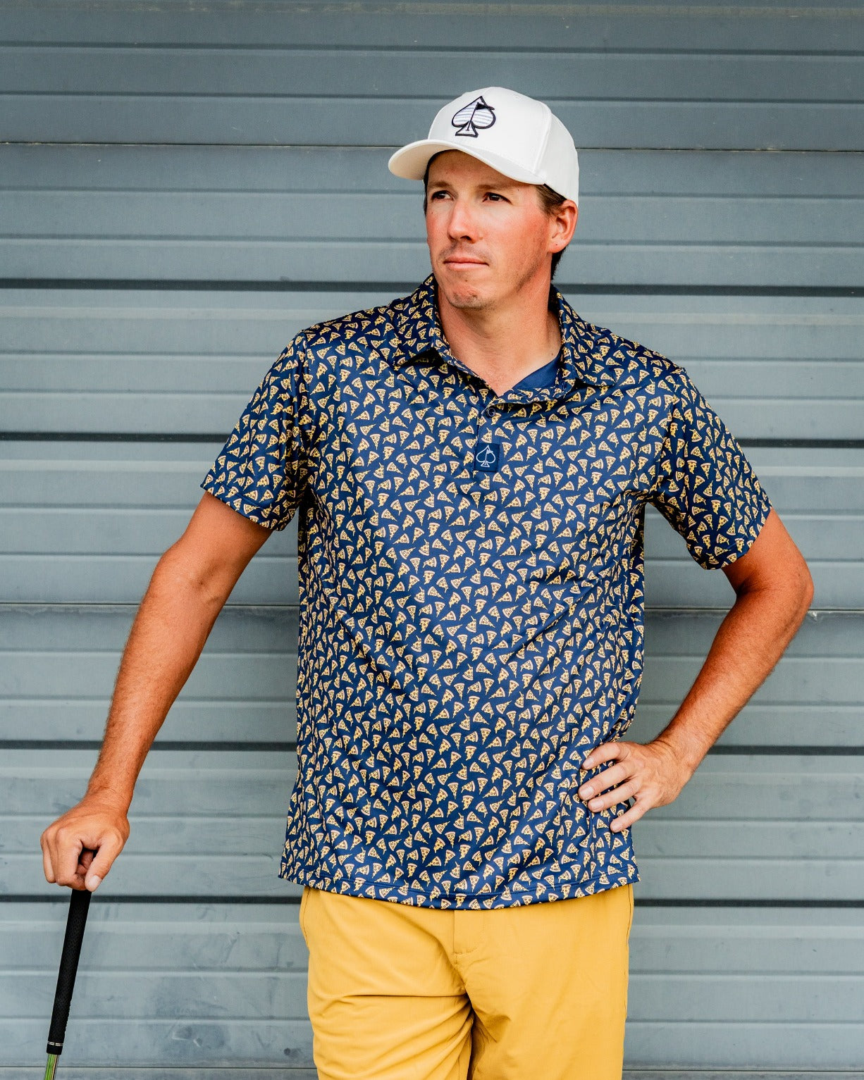 A golfer wearing a patterned navy polo shirt and yellow shorts stands confidently with a golf club, against a gray backdrop.