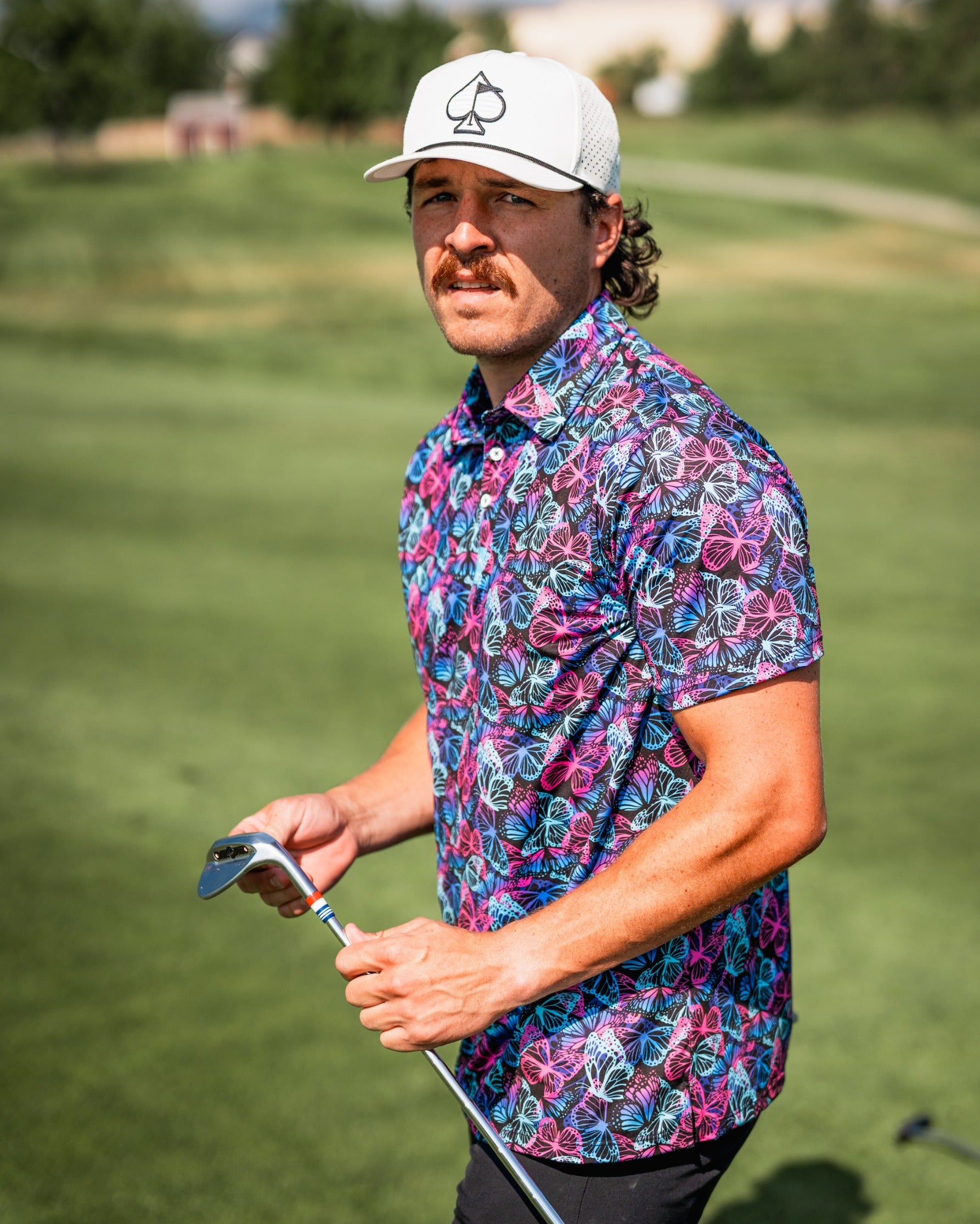A golfer stands on the course wearing a vibrant Electric Butterfly shirt, holding a golf club, showcasing bold floral patterns.