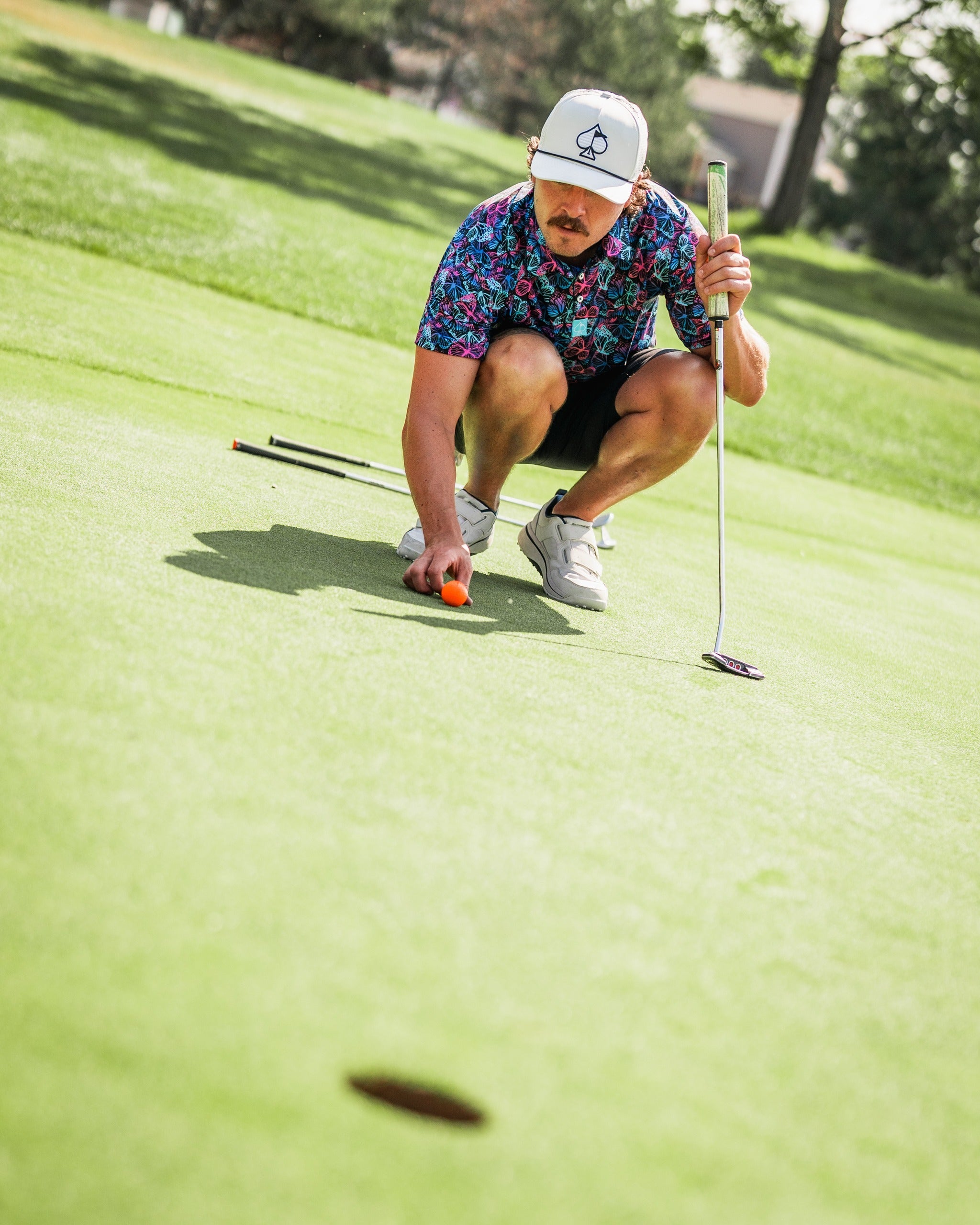 A golfer in a colorful floral shirt kneels on the green, preparing to putt a ball into the hole.