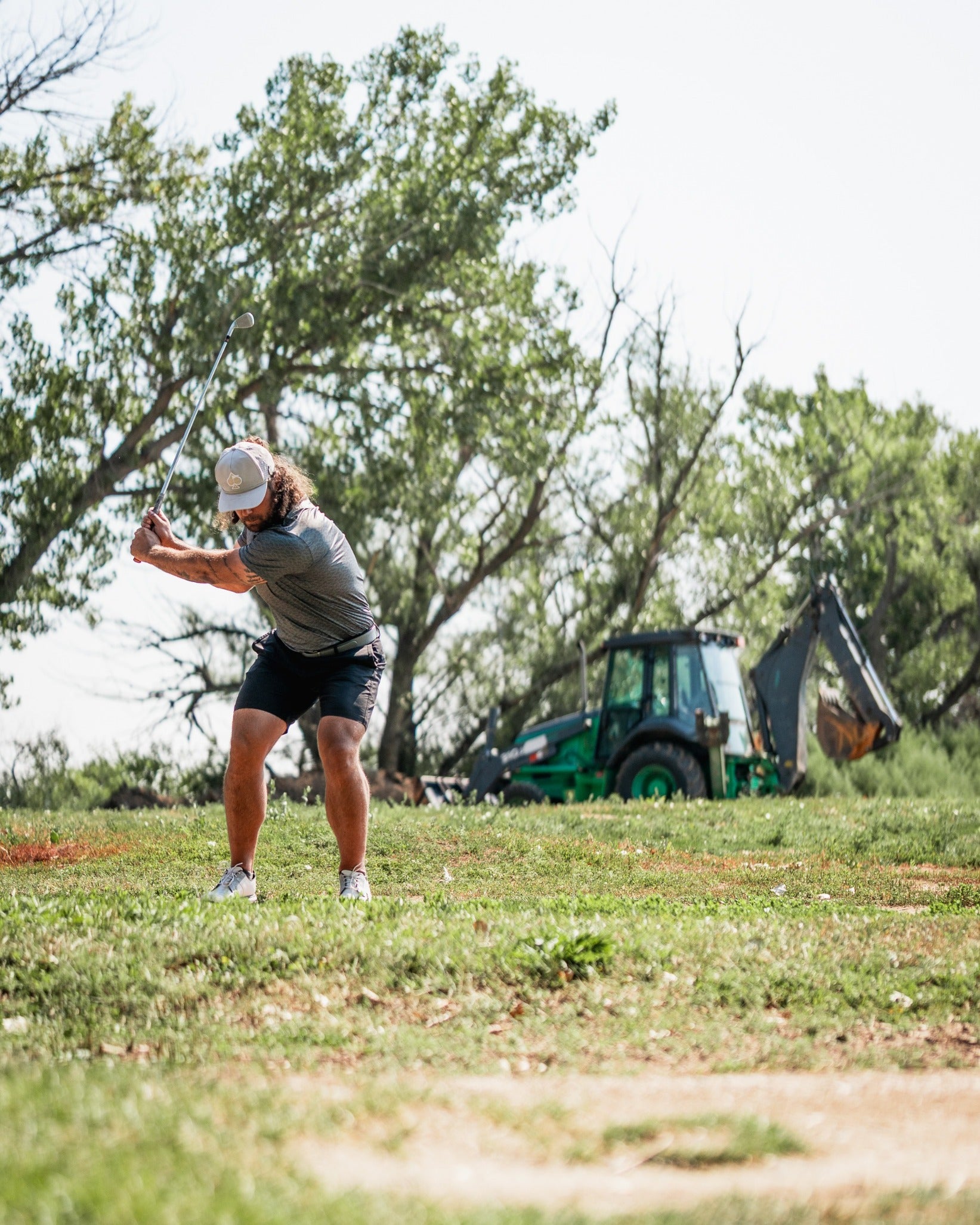 Golfer in a gray shirt and shorts swings a club on a course with a tractor in the background, showcasing the Large Mouth headcover.