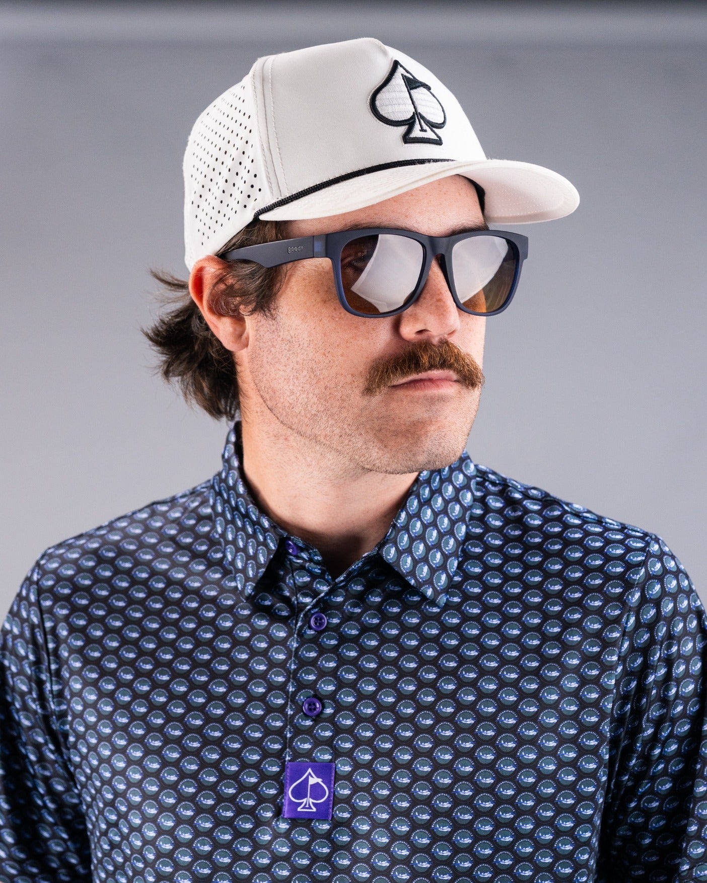 Man wearing a patterned golf shirt and a white cap with a logo, showcasing sunglasses and a stylish look for golfers.