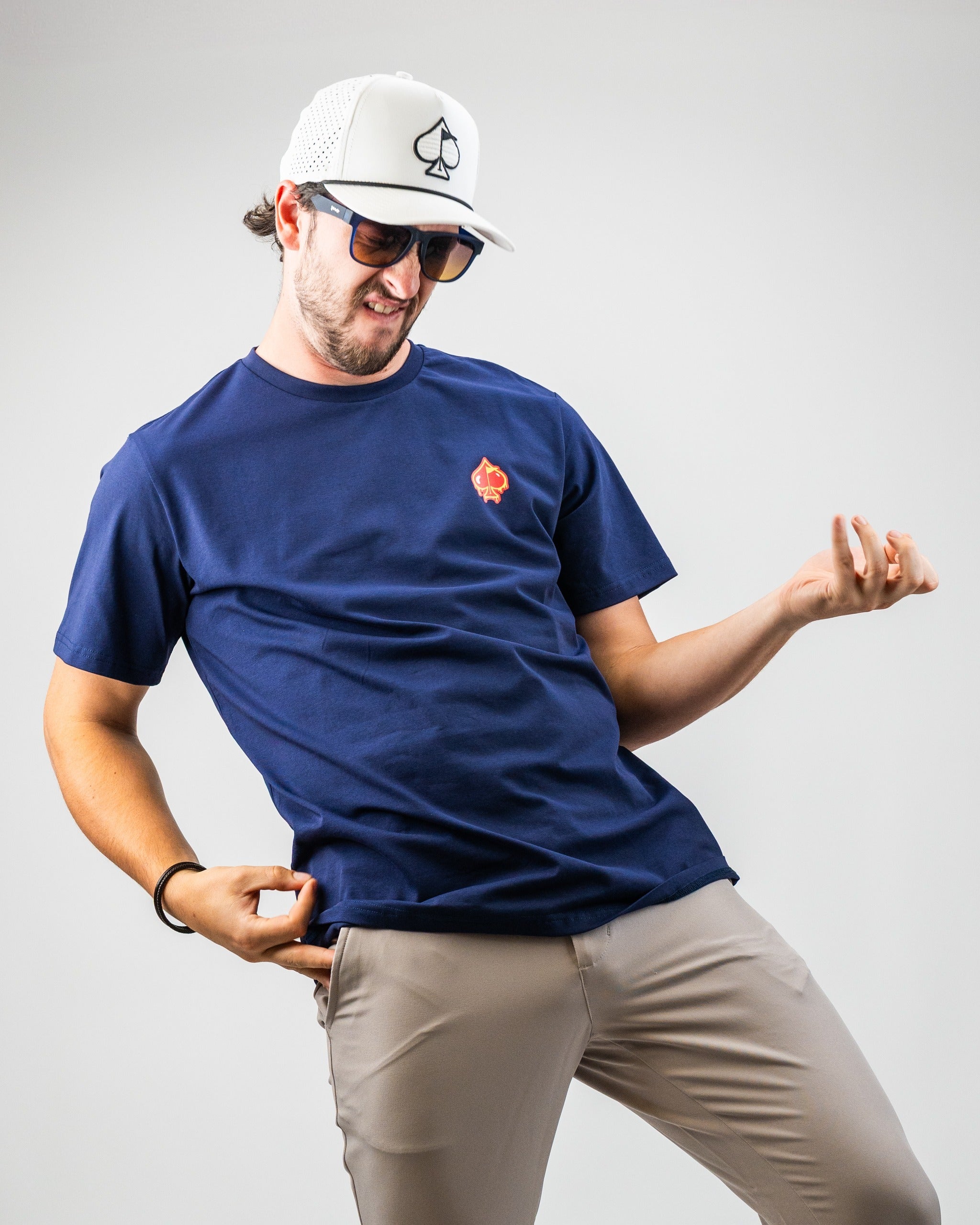 Man wearing a navy golf T-shirt with a red spade logo, beige pants, white cap with spade emblem, and sunglasses, striking ...