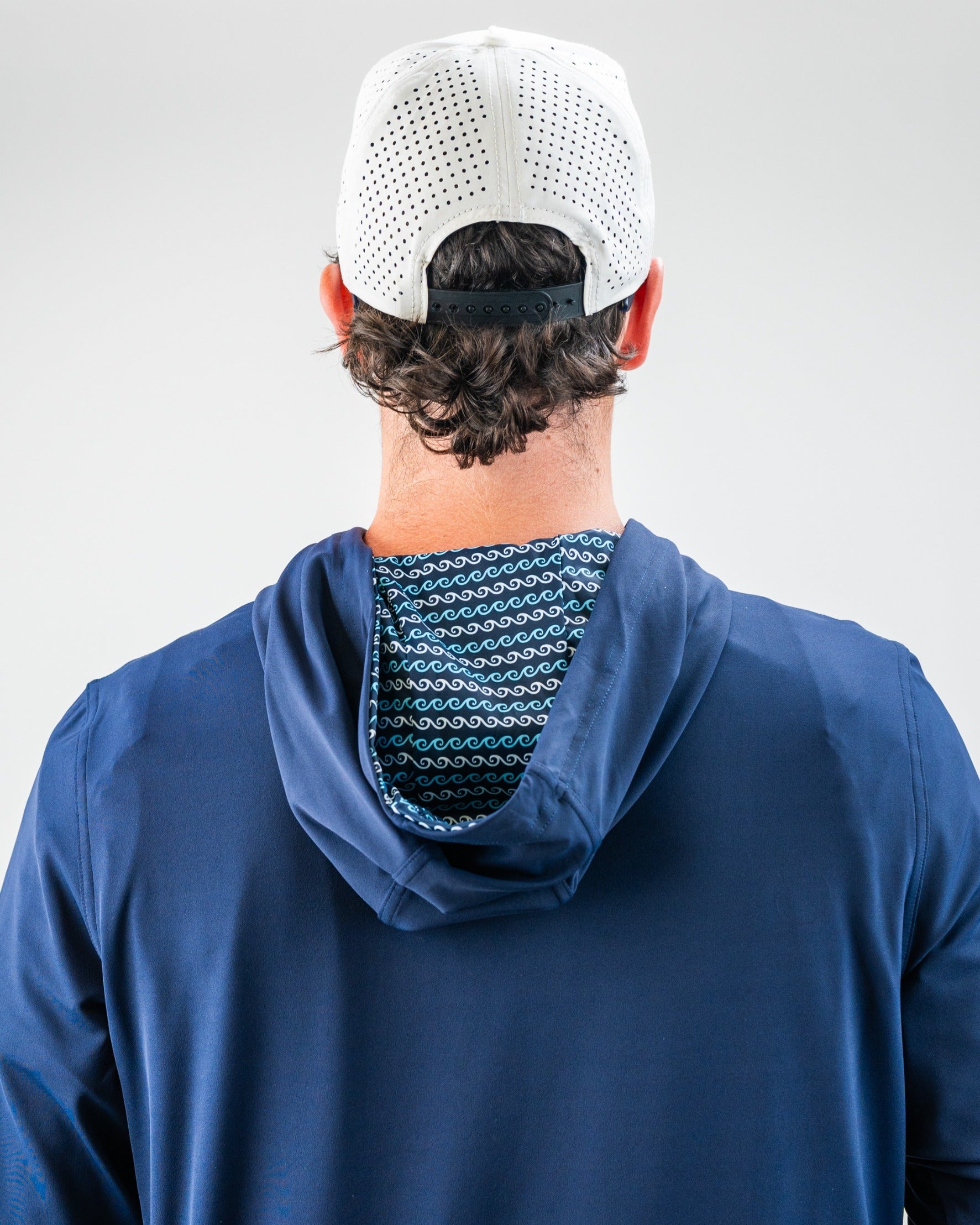 Back view of a golfer wearing a white perforated cap, navy blue jacket, and a patterned hood with waves in blue, black, an...