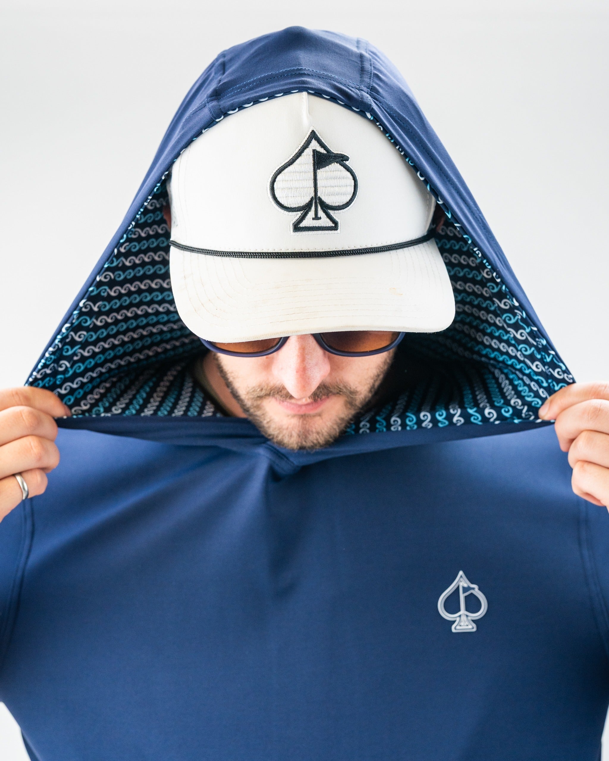 Man wearing a navy blue jacket with a spade logo, removing the hood, with a white cap featuring a spade and flag embroider...