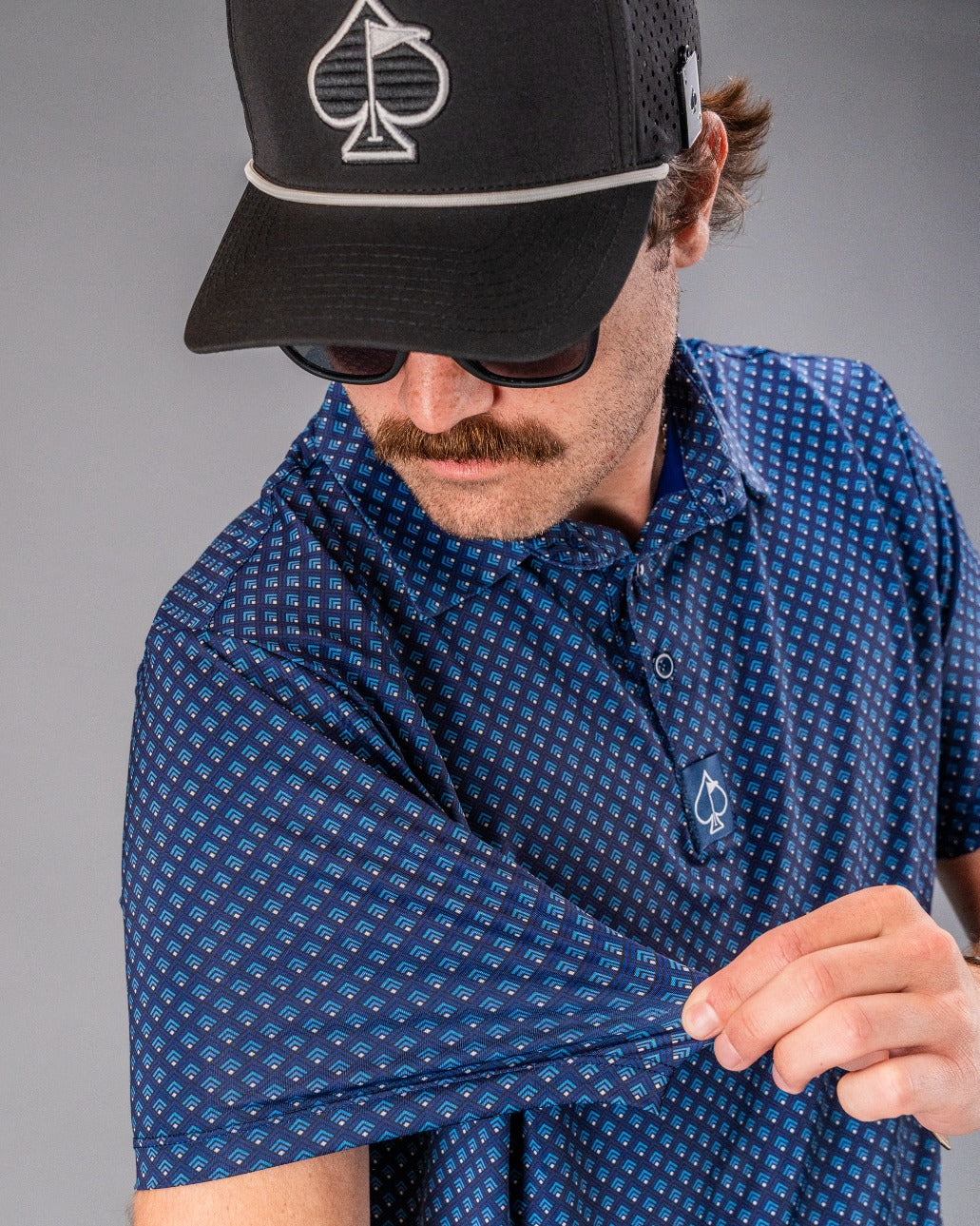 Man wearing a black cap with spade logo, sunglasses, and a blue patterned golf shirt, showcasing bold golf apparel.