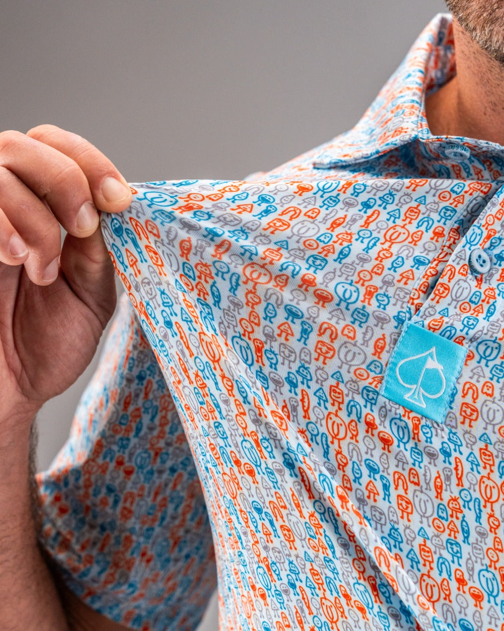 Close-up of a men's golf shirt with a colorful, playful print in blue, orange, gray, and white, featuring various cartoon ...