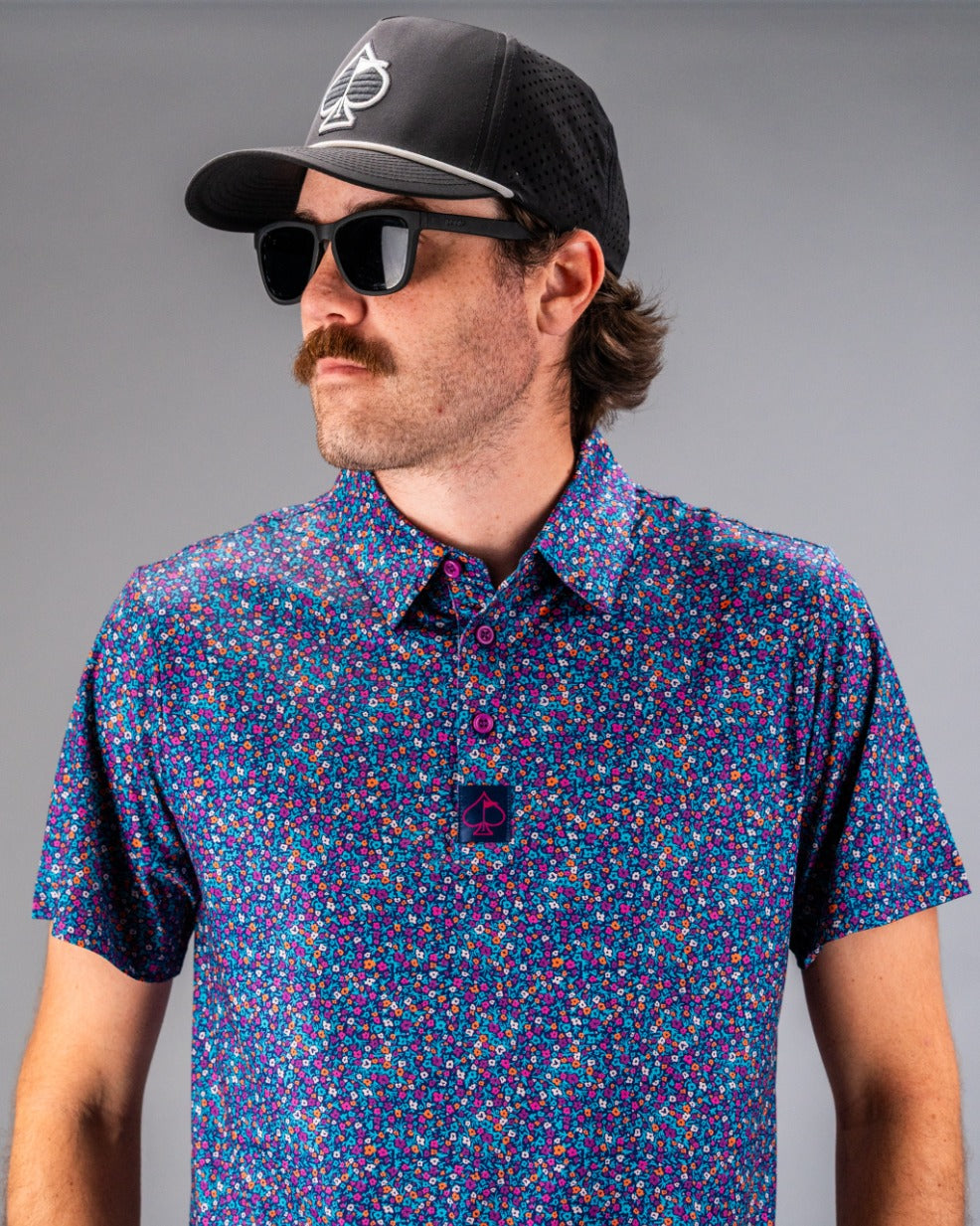 Man wearing a vibrant lavender floral golf polo shirt, paired with a black cap and sunglasses, showcasing a bold style.