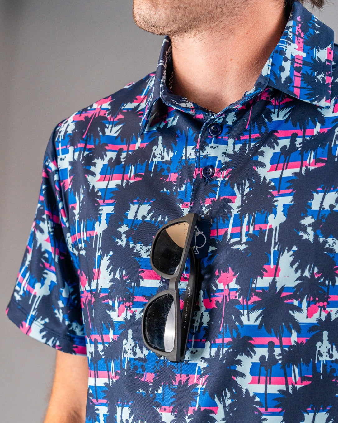 Close-up of a golfer wearing a bold, colorful Hawaiian shirt with palm trees and sunset stripes, accessorized with black s...