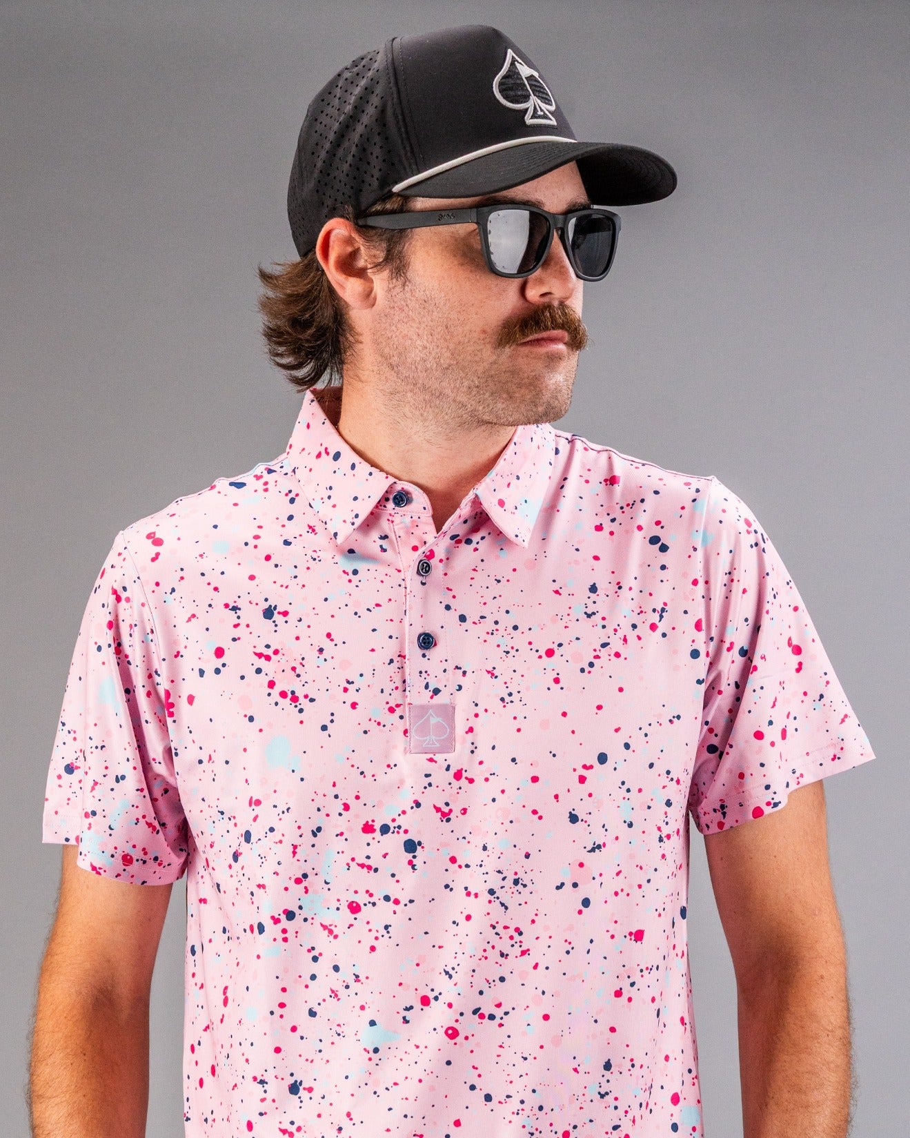 Men's pink splatter golf polo featuring vibrant paint splashes, paired with a black cap and sunglasses for a bold look.