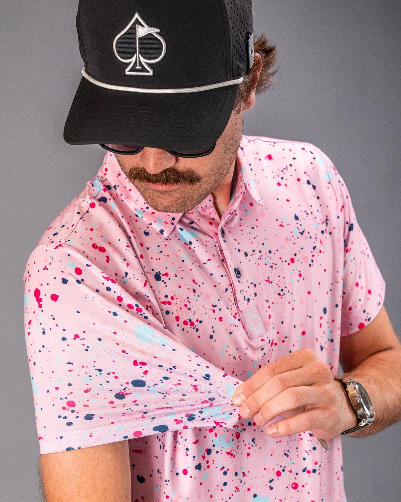 A golfer showcases a pink splatter polo shirt with vibrant paint-like patterns, paired with a black cap and sunglasses.